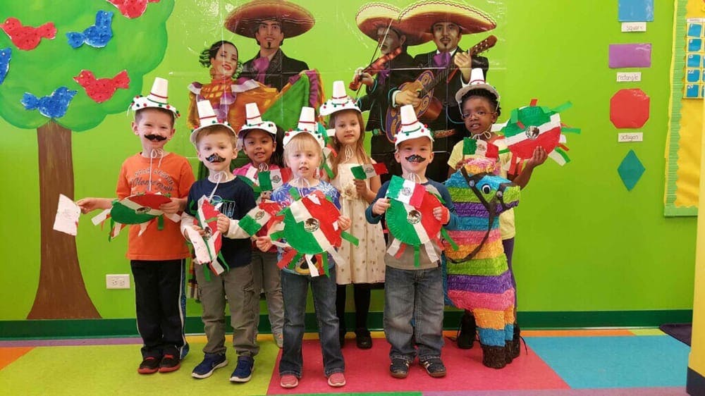 Children in paper hats and drawn-on mustaches holding Mexican flag crafts and piñata decorations