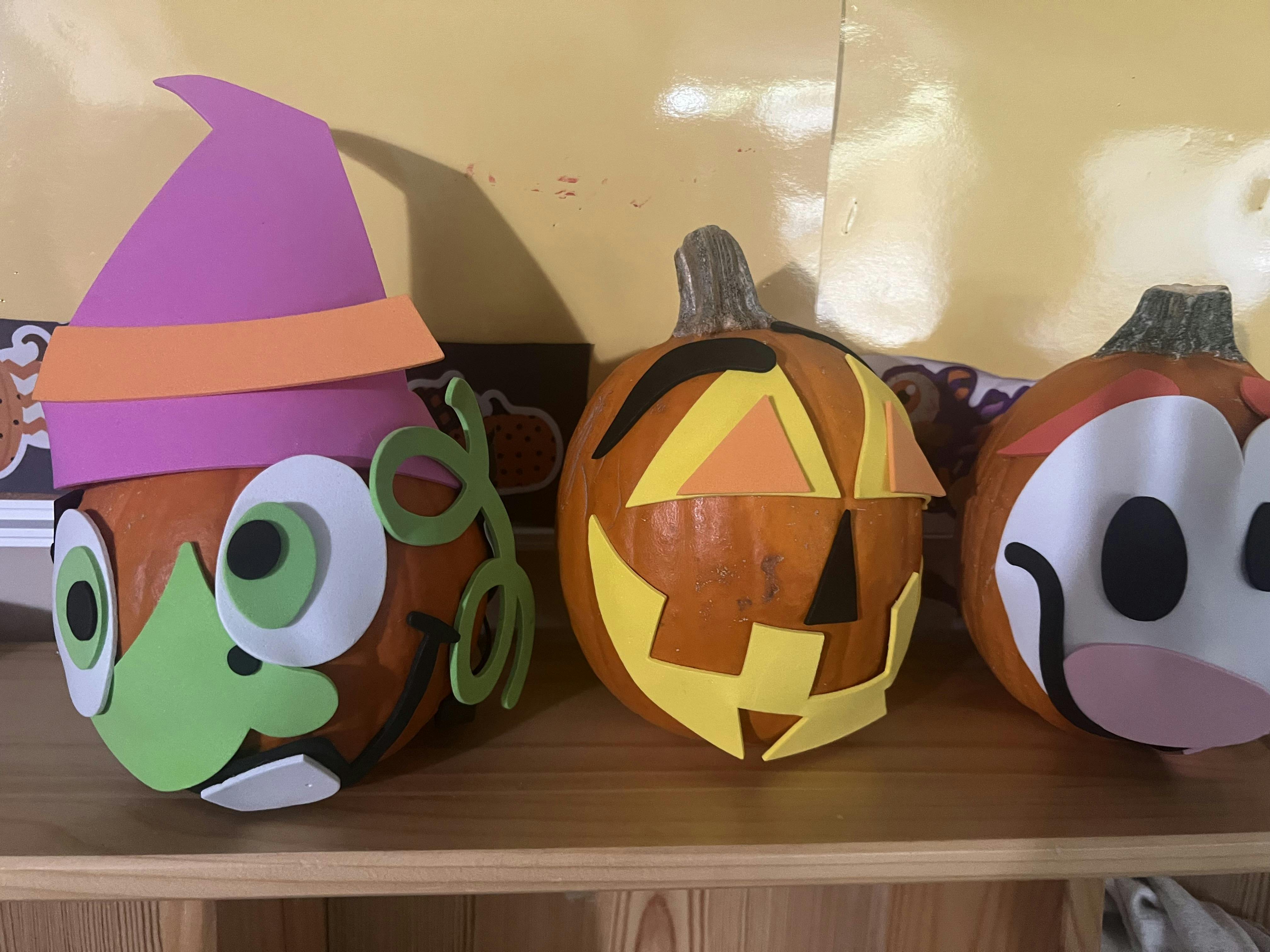 Three pumpkins decorated with foam craft shapes on a wooden shelf