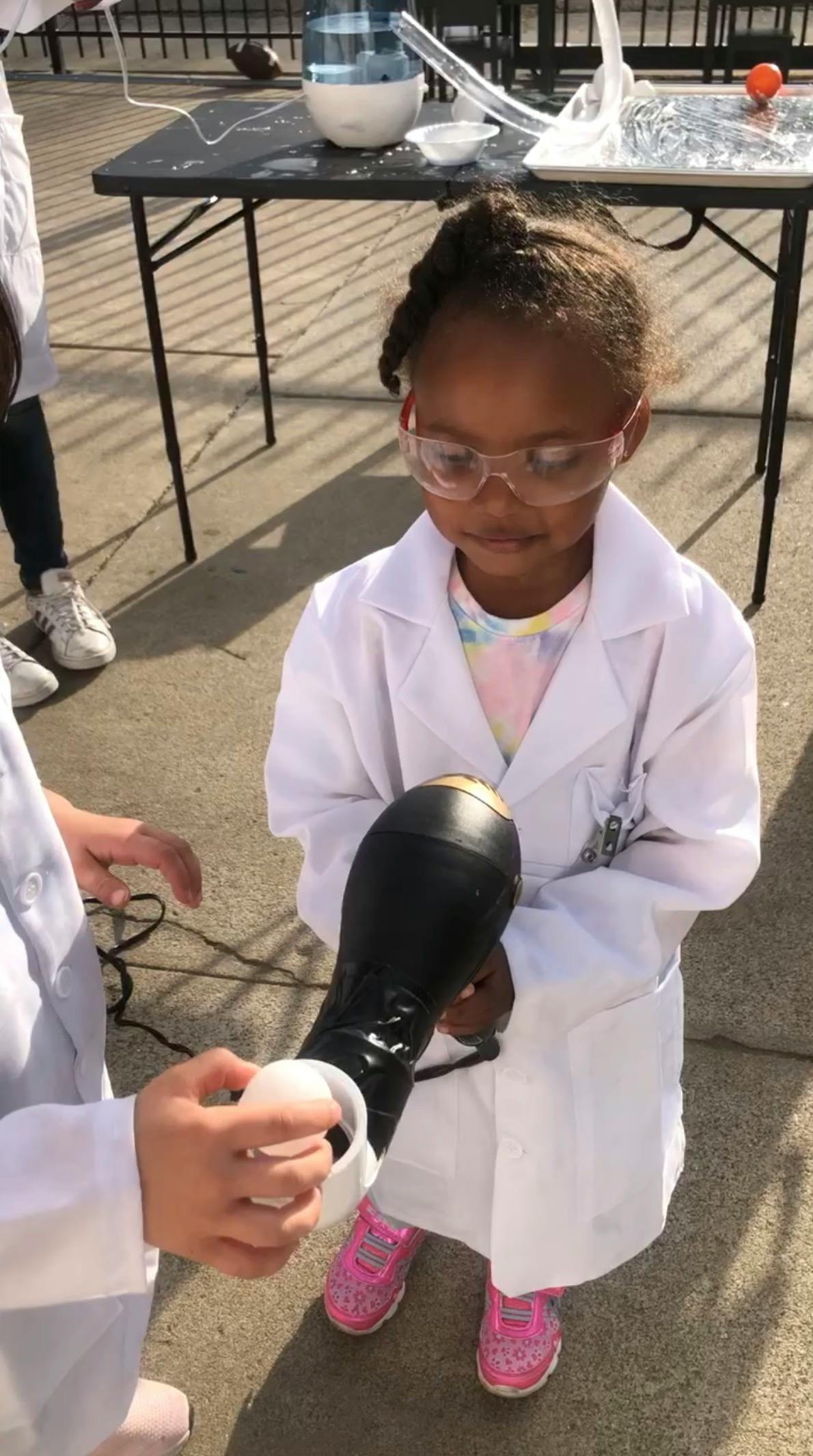Child wearing a lab coat and safety goggles holds a hair dryer