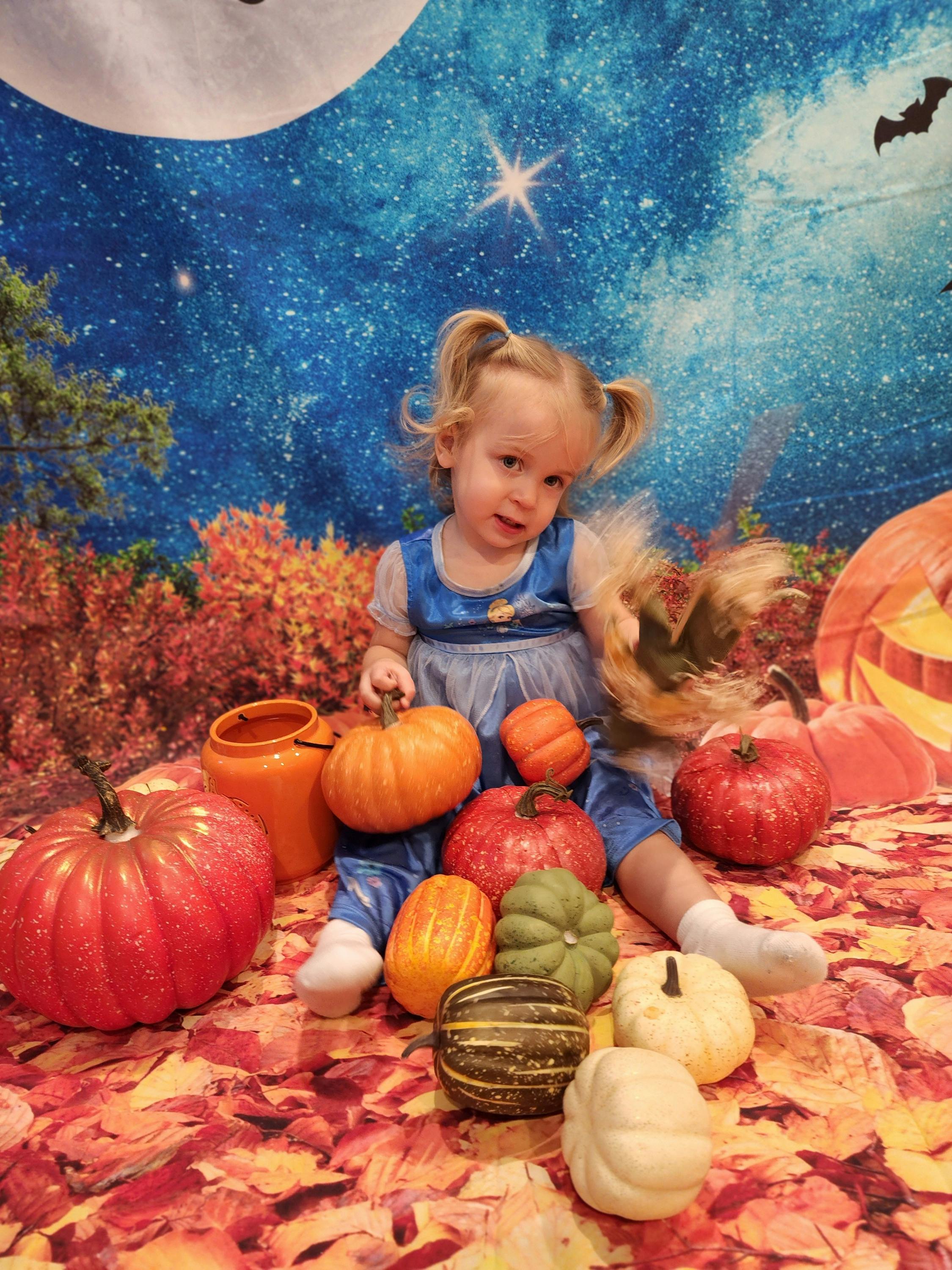 Child in a blue dress sitting with pumpkins and gourds on a fall-themed backdrop