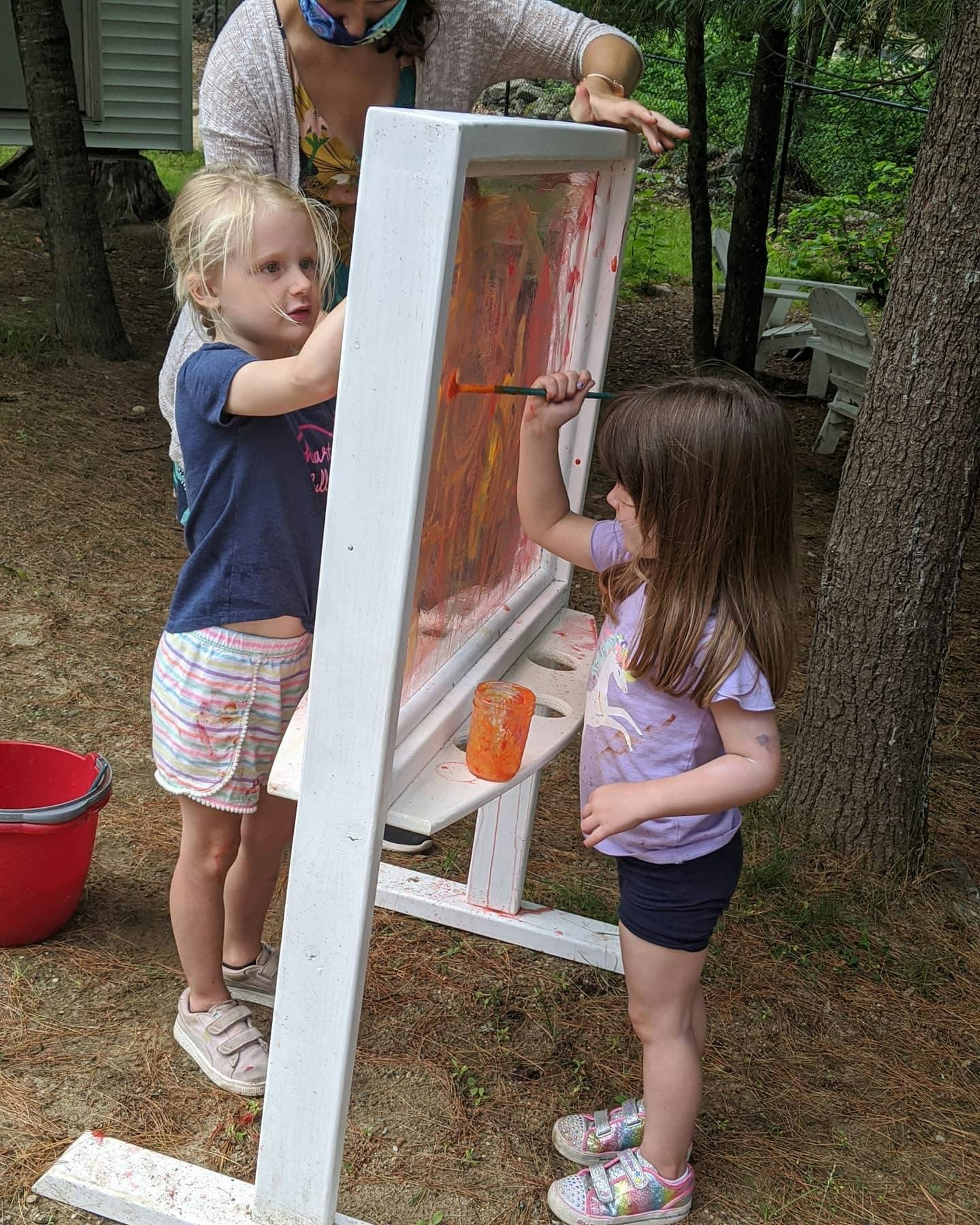 Two children painting on an easel outdoors with an adult nearby