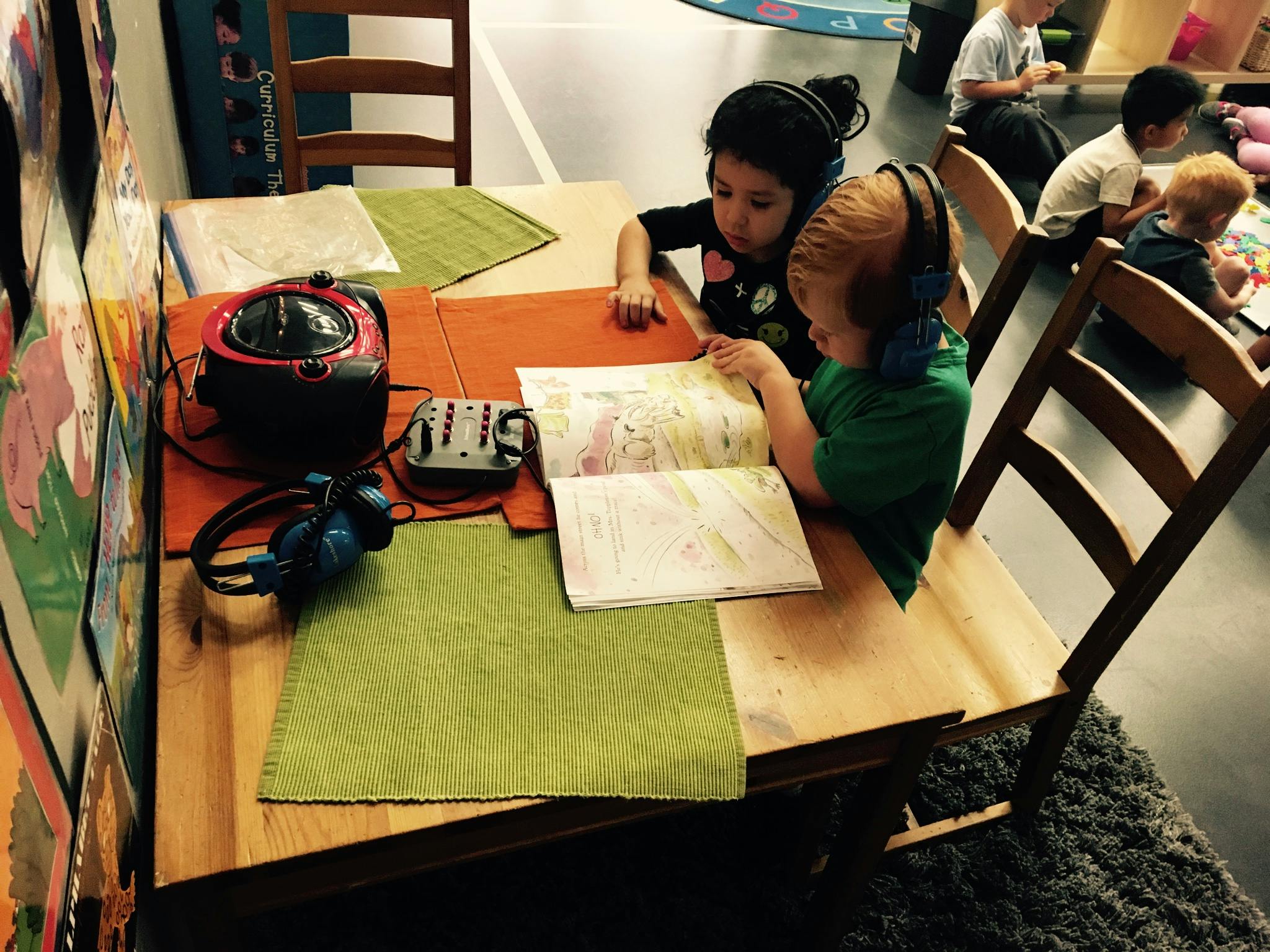 Two children with headphones reading a book at a table