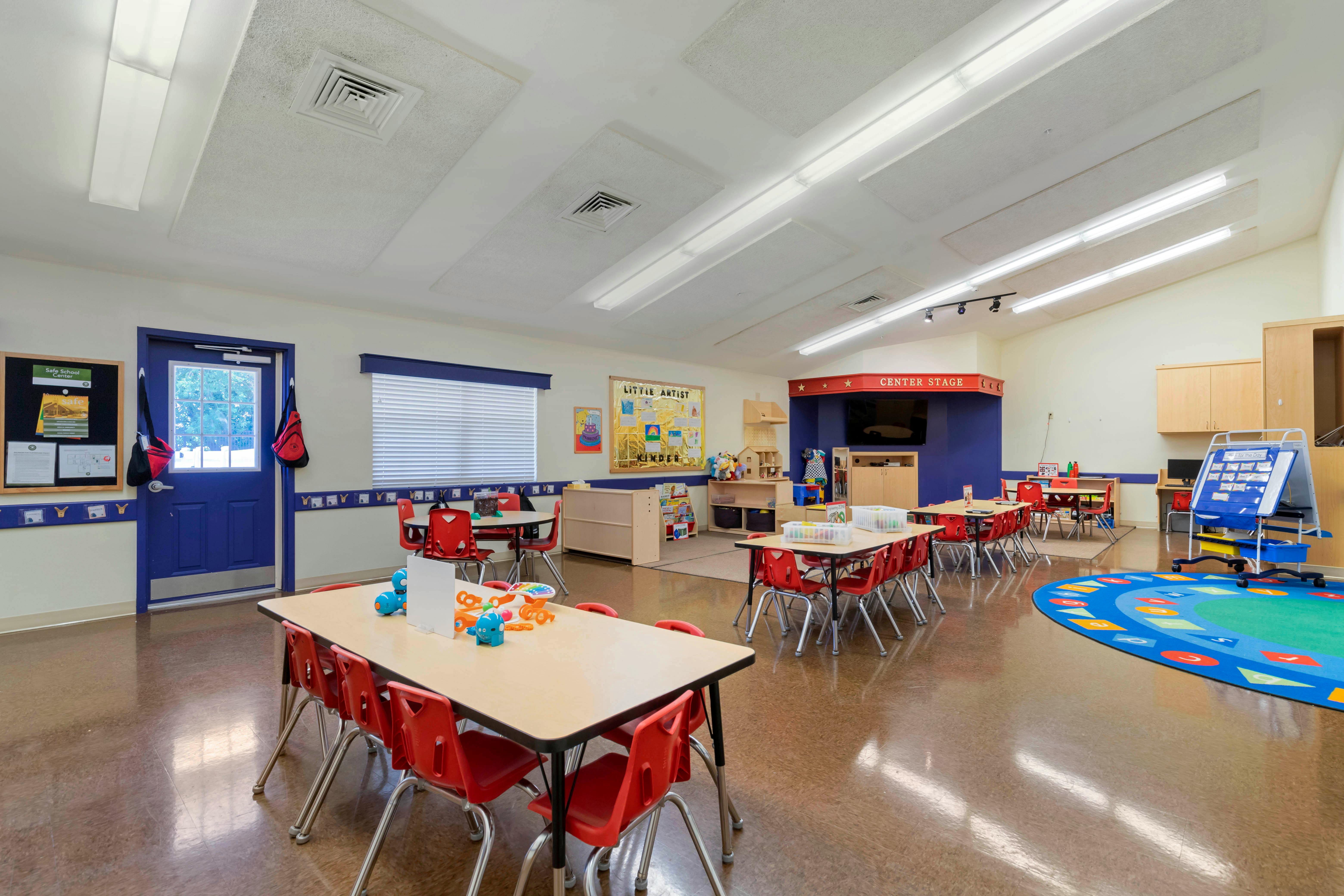 Classroom with small tables and red chairs, a blue stage area, and a colorful rug