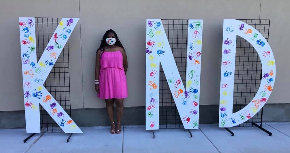 Person in a pink dress standing next to large letters spelling KIND