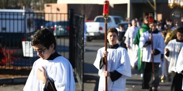 People in white robes walking outdoors, one holding a staff with a red top