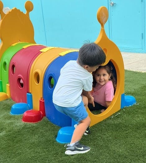 Two children playing in a colorful caterpillar tunnel on green turf