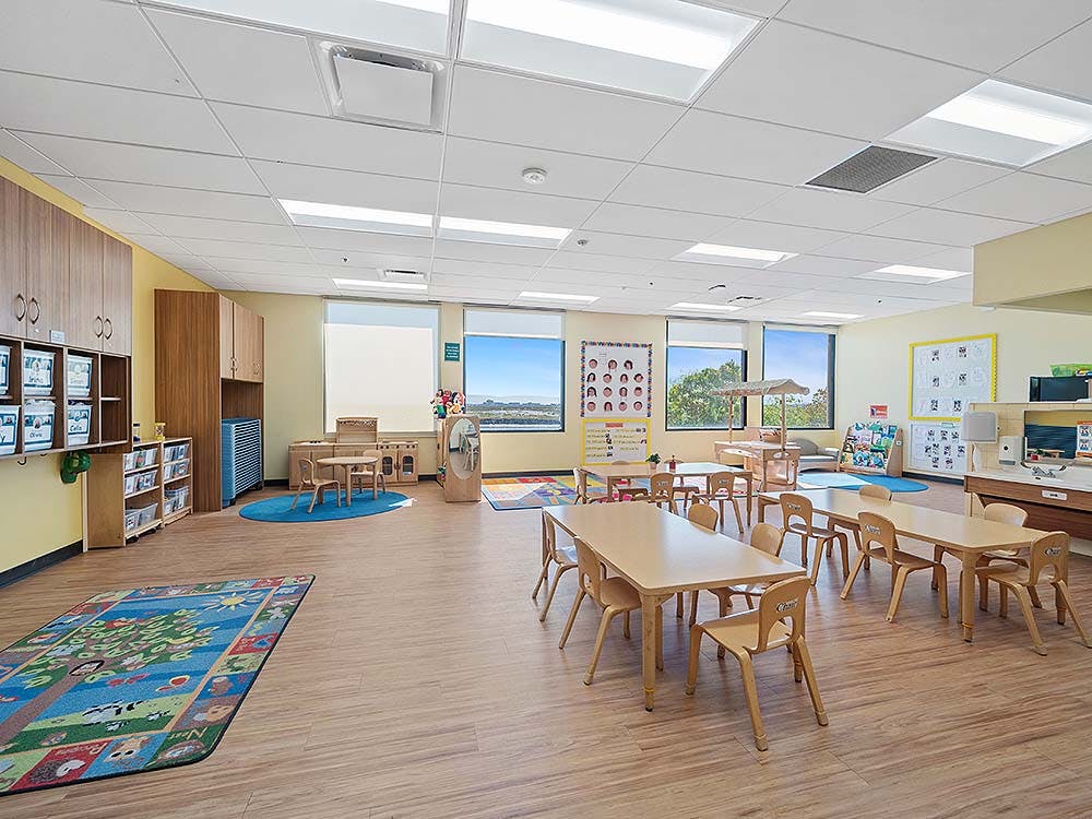 Empty classroom with small tables and chairs, a play rug, and large windows