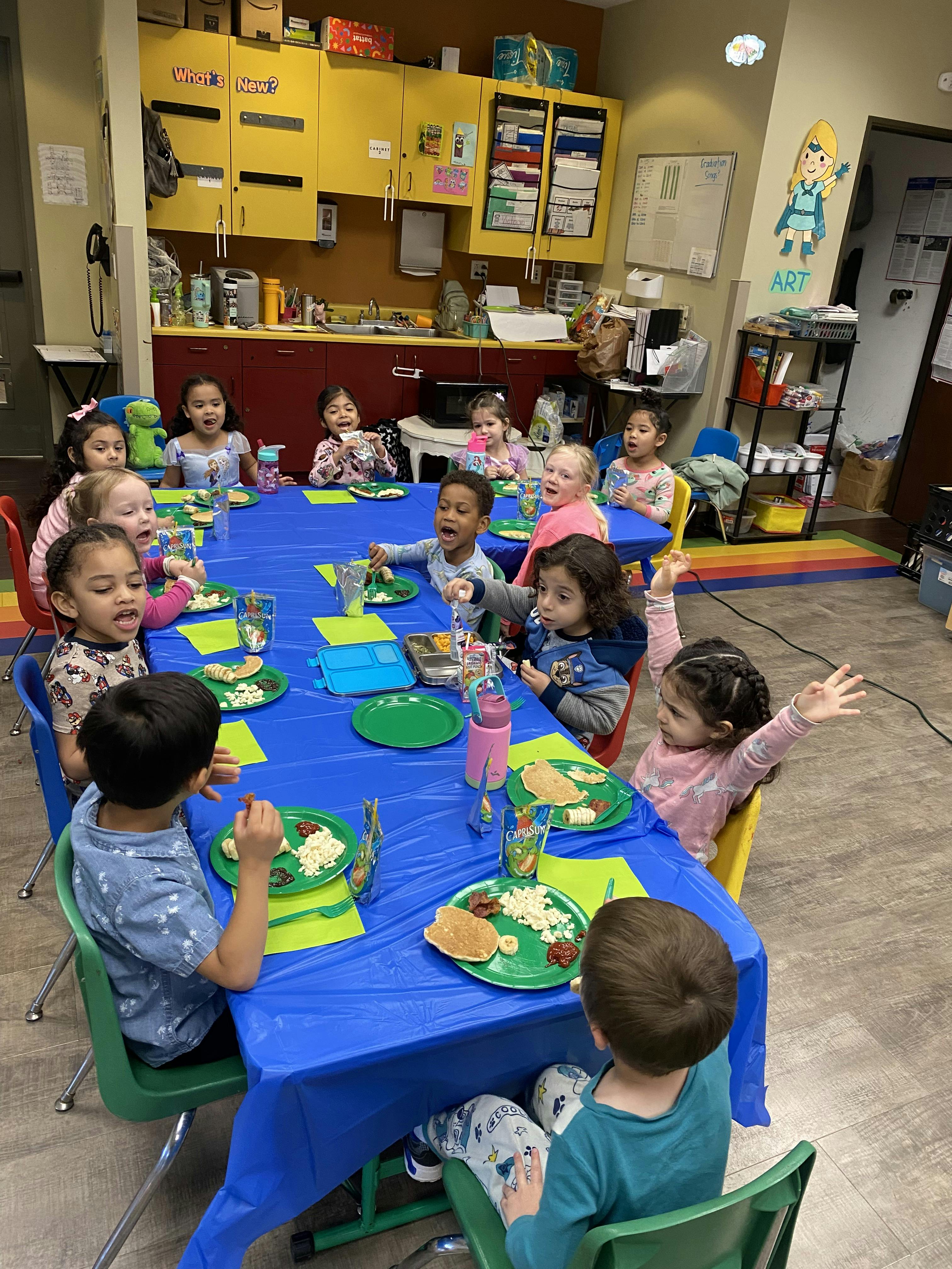 Group of children eating lunch at a large blue table in a classroom