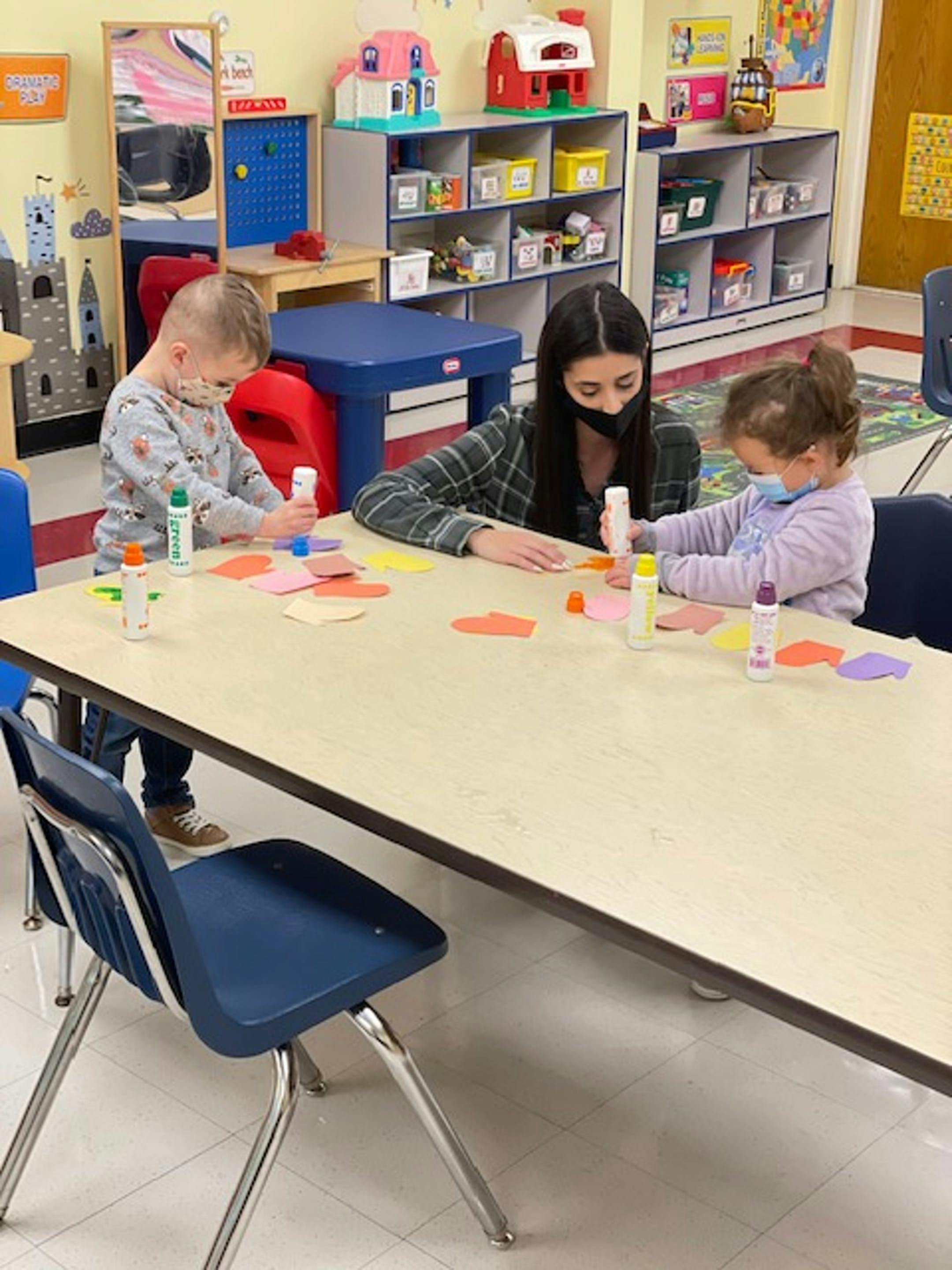 Adult and two children in masks doing crafts at a table in a classroom