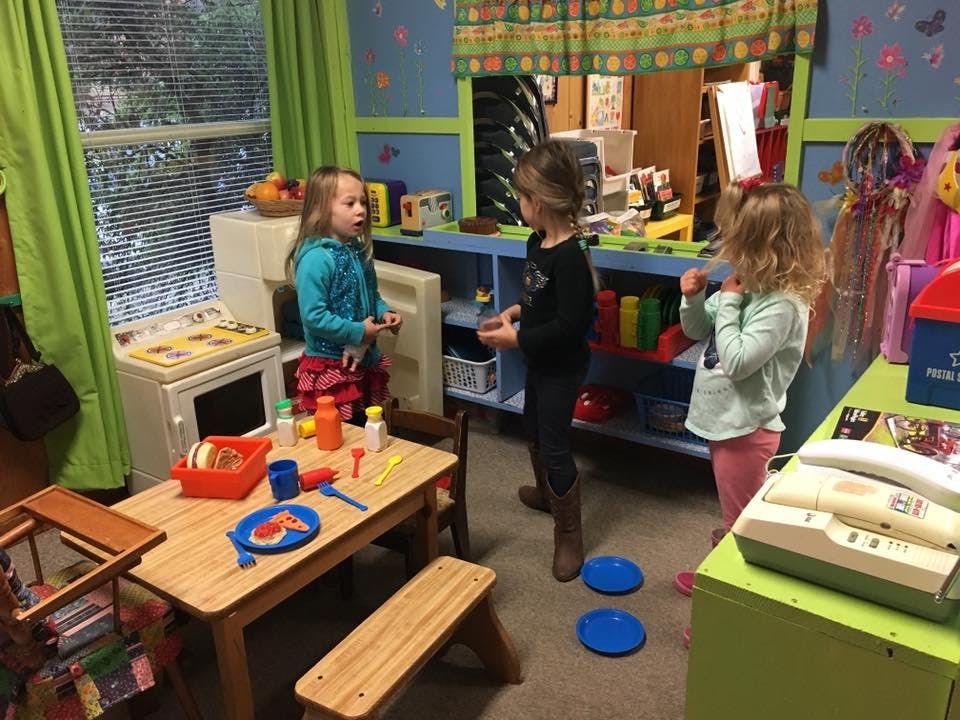 Three children playing in a classroom with a play kitchen and craft area