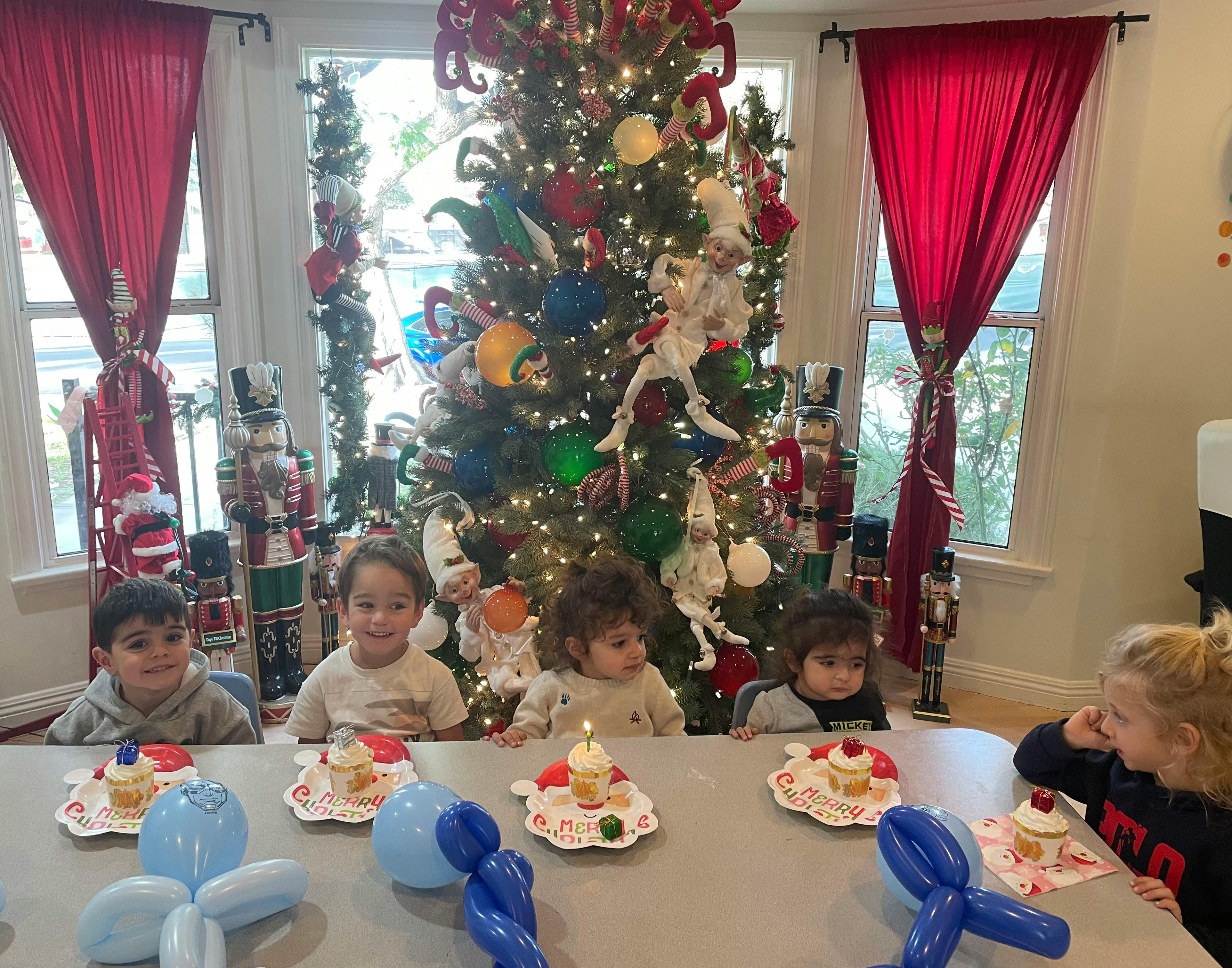 Children sitting at a table with cupcakes and balloons in front of a Christmas tree