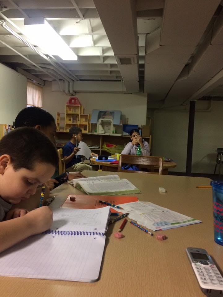 Child writing in a notebook at a table with other people in the background