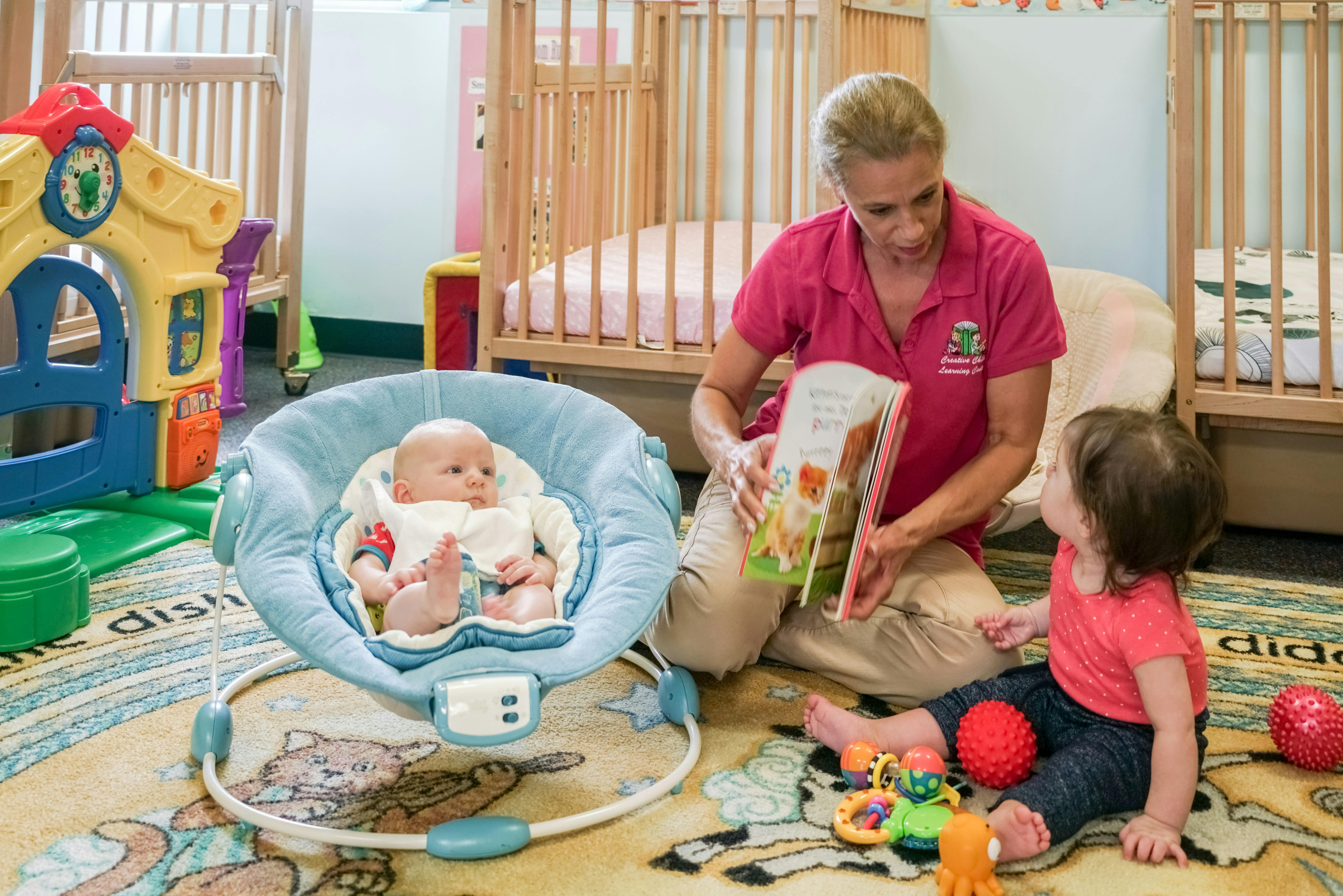 Adult reading a book to a child, with an infant in a bouncer nearby