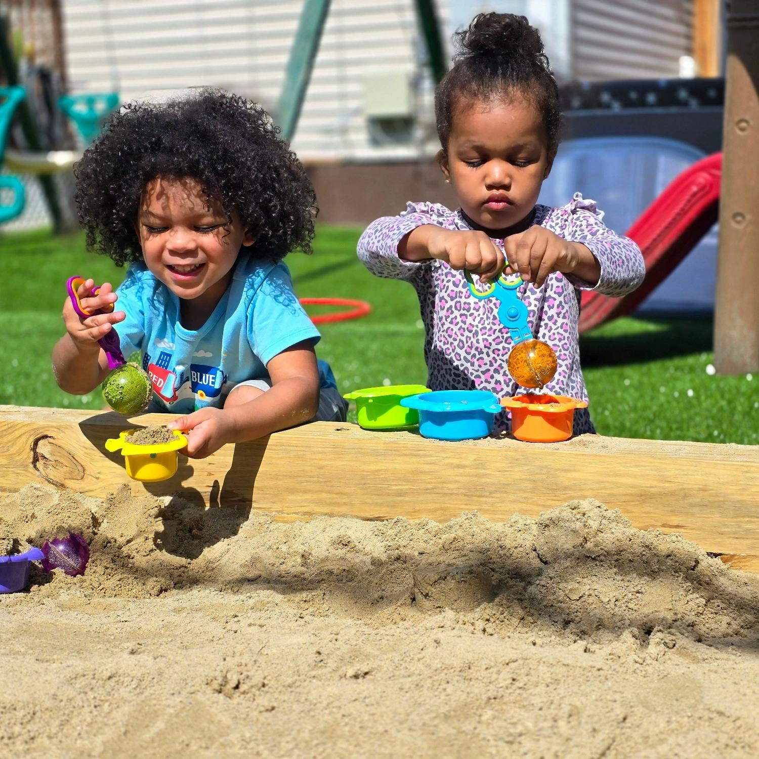 Two children playing with sand and plastic tools in a sandbox