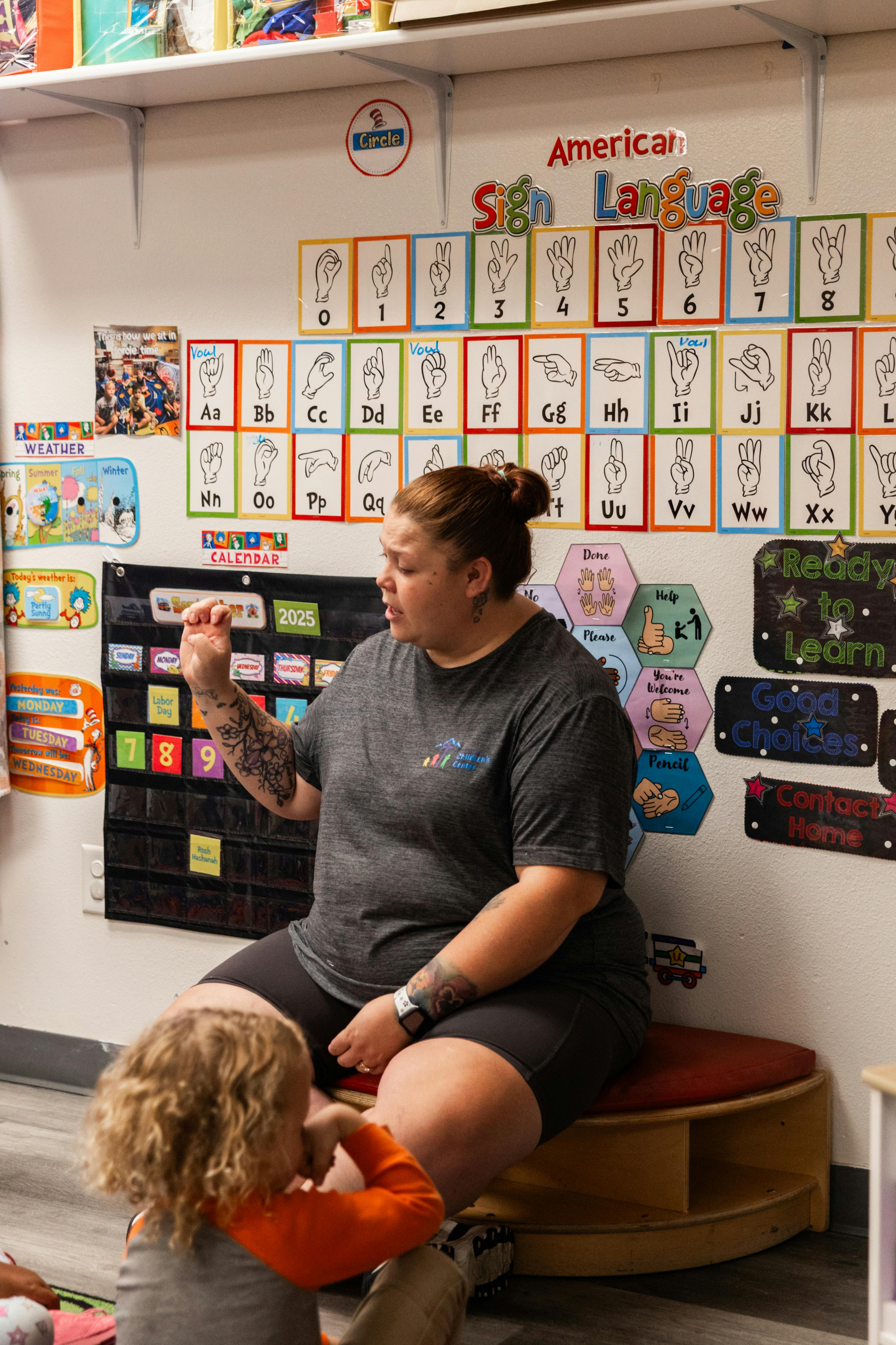 Adult talking to child in a classroom with ASL alphabet chart on wall