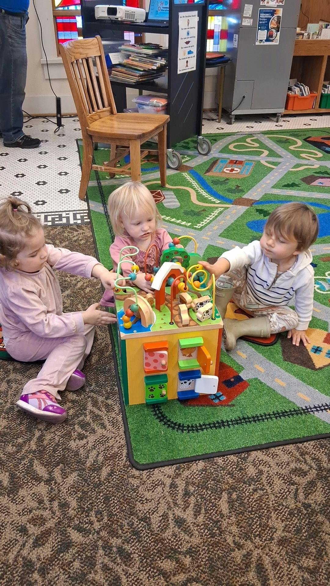 Three children playing with a large wooden activity cube on a rug