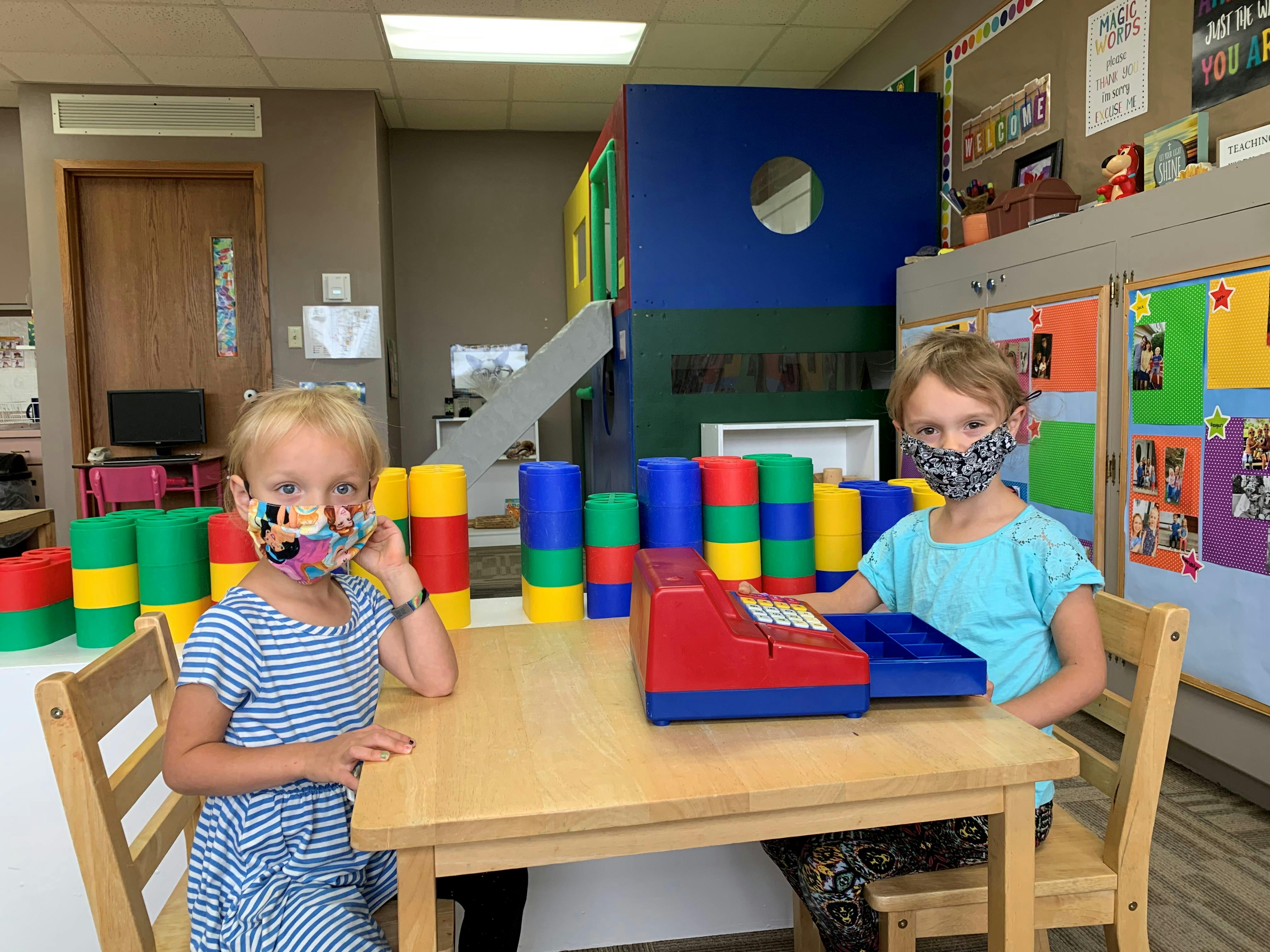 Two children wearing masks sitting at a table with colorful blocks and a toy cash register