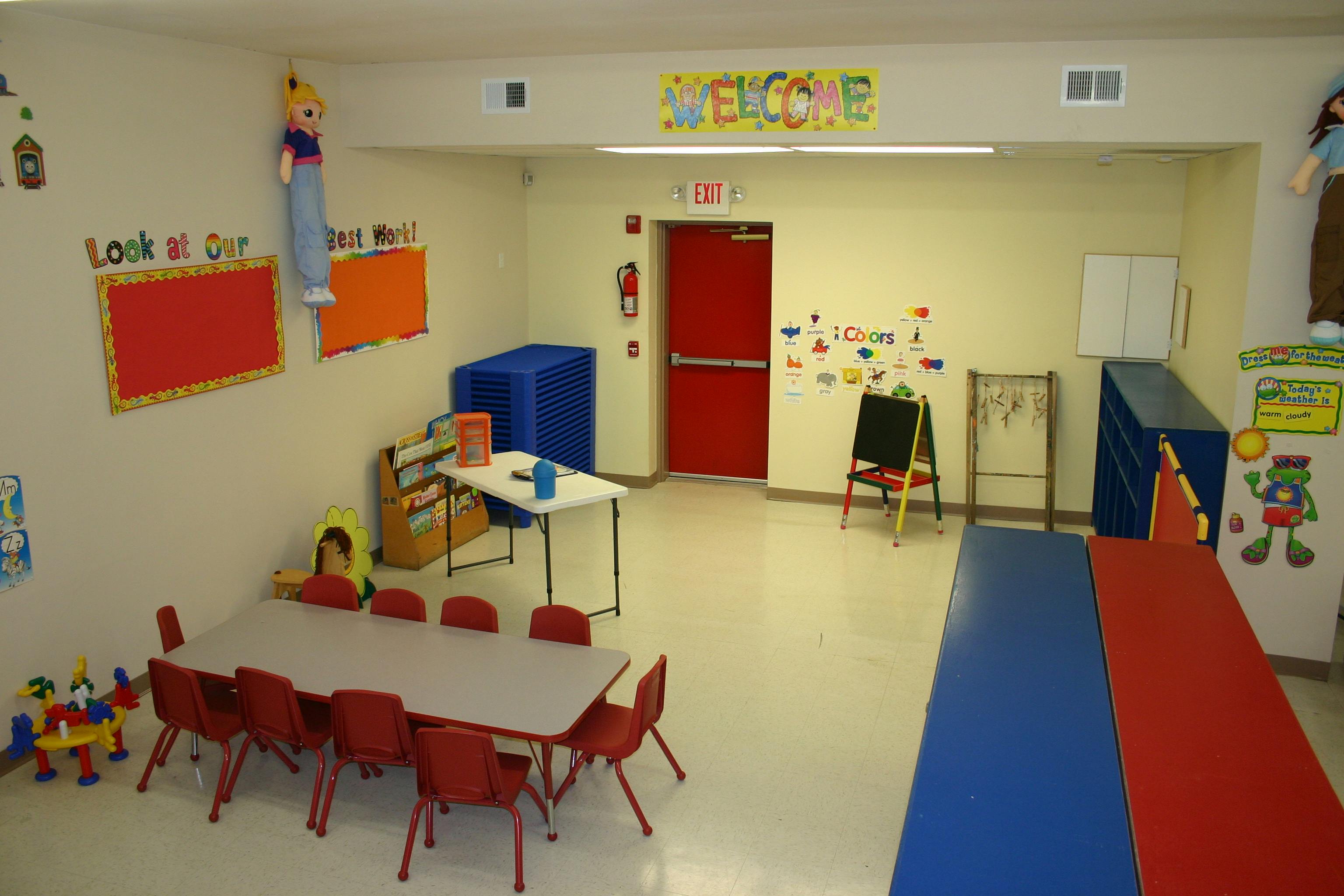 Empty classroom with small tables and chairs, red door, and colorful decorations