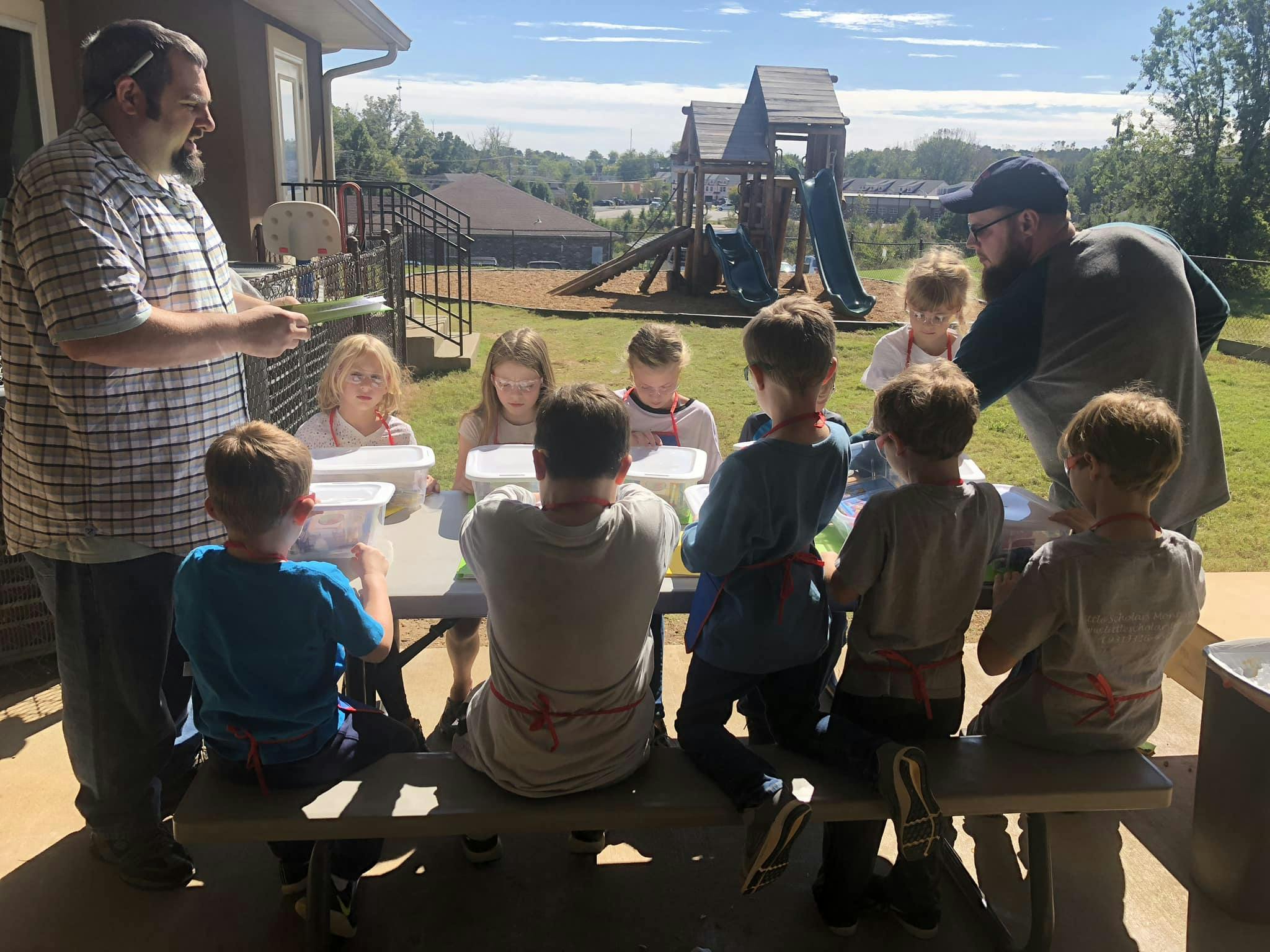 Two adults interacting with a group of children seated at a table outdoors