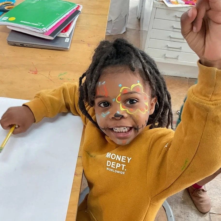 Child with face paint smiling and holding a pencil at a table