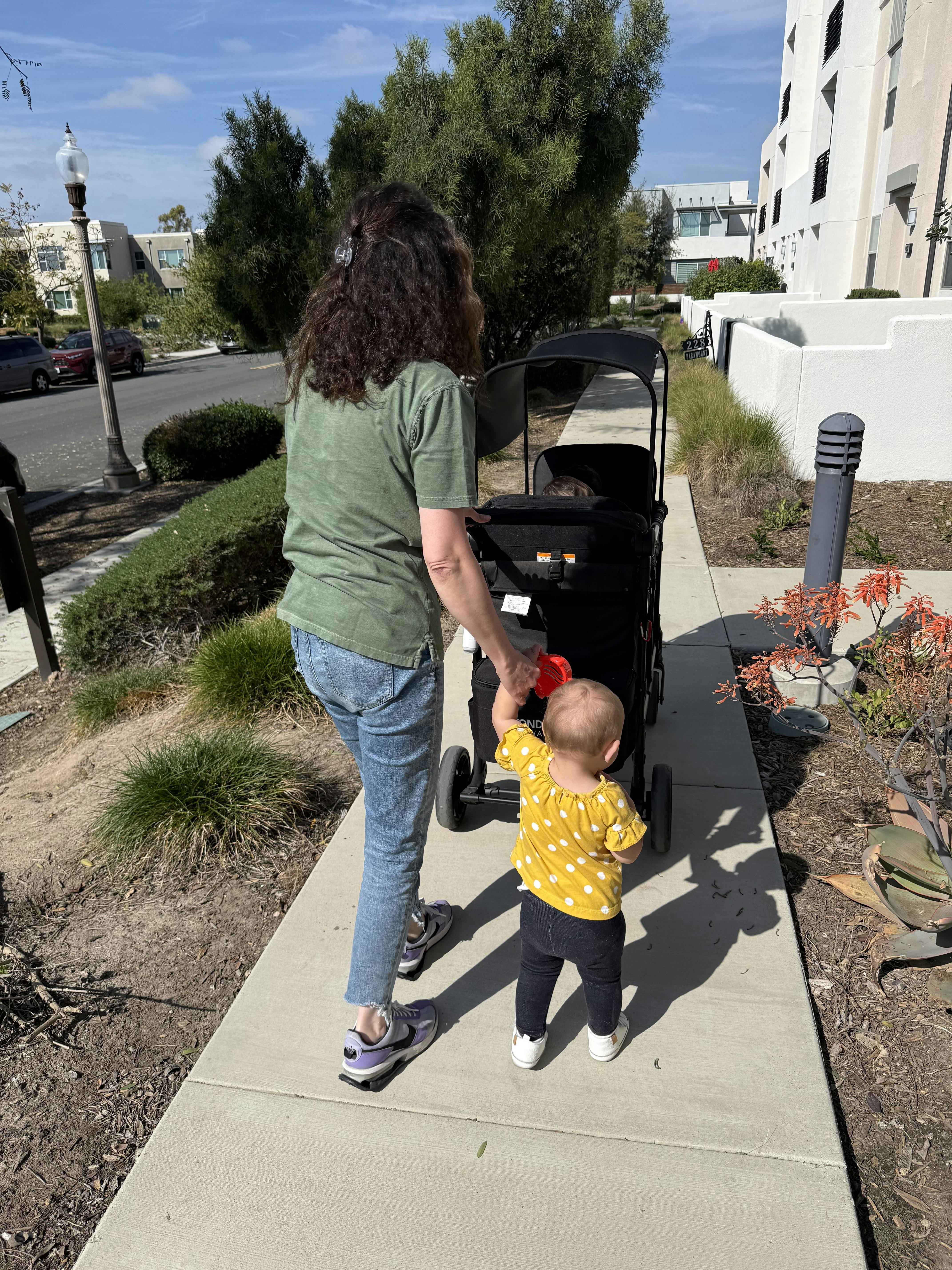 Person pushing a stroller with a child walking beside it on a sidewalk