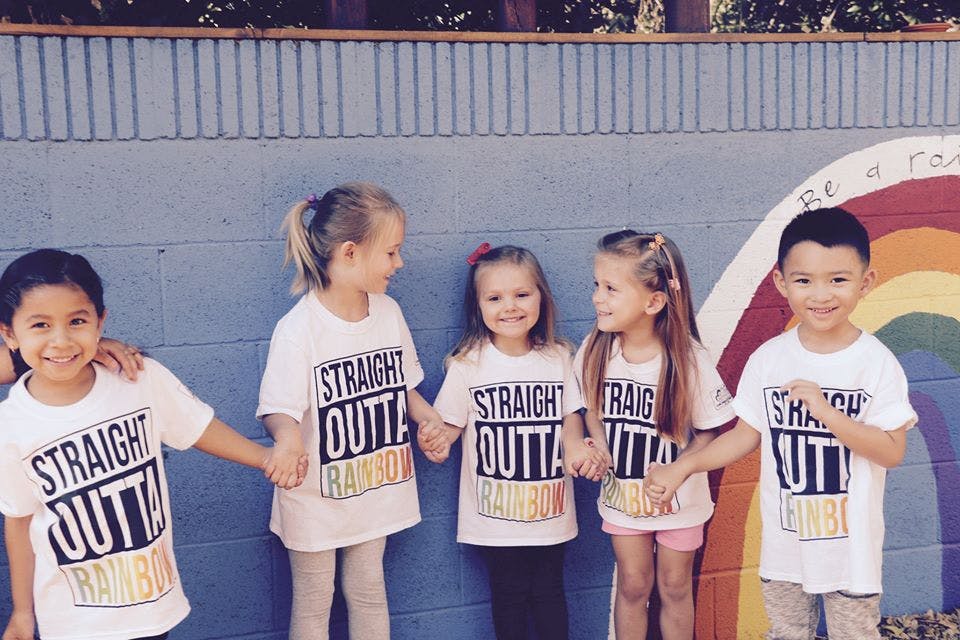 Five children holding hands in front of a wall with a rainbow painted on it