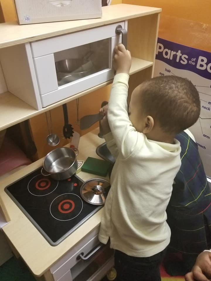 Child playing with a toy kitchen set, reaching for the microwave door
