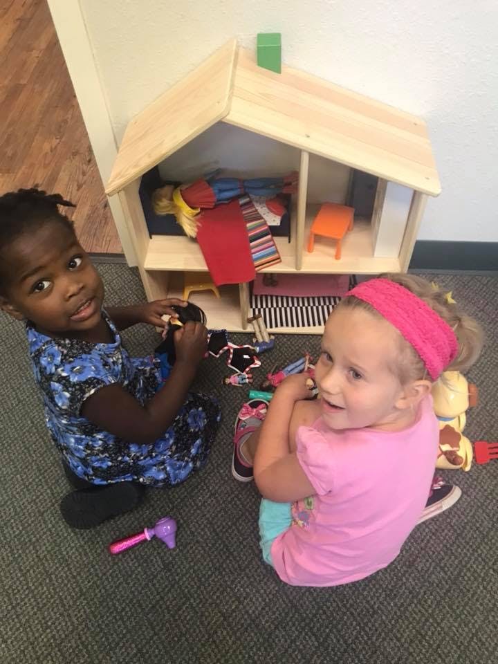 Two children playing with a dollhouse on the floor next to a wooden dollhouse