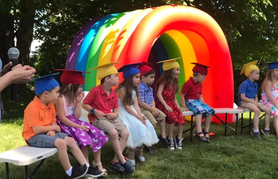 Children in graduation caps sitting on benches in front of a rainbow arch