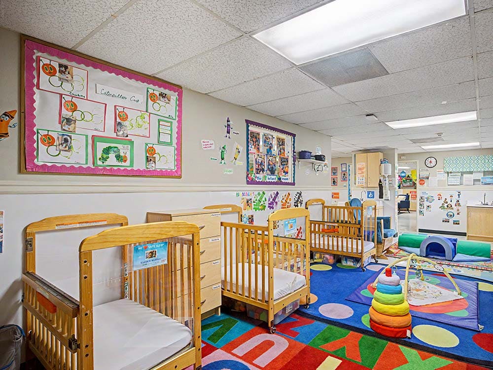 cribs in a brightly lit daycare room with colorful rugs and decorations