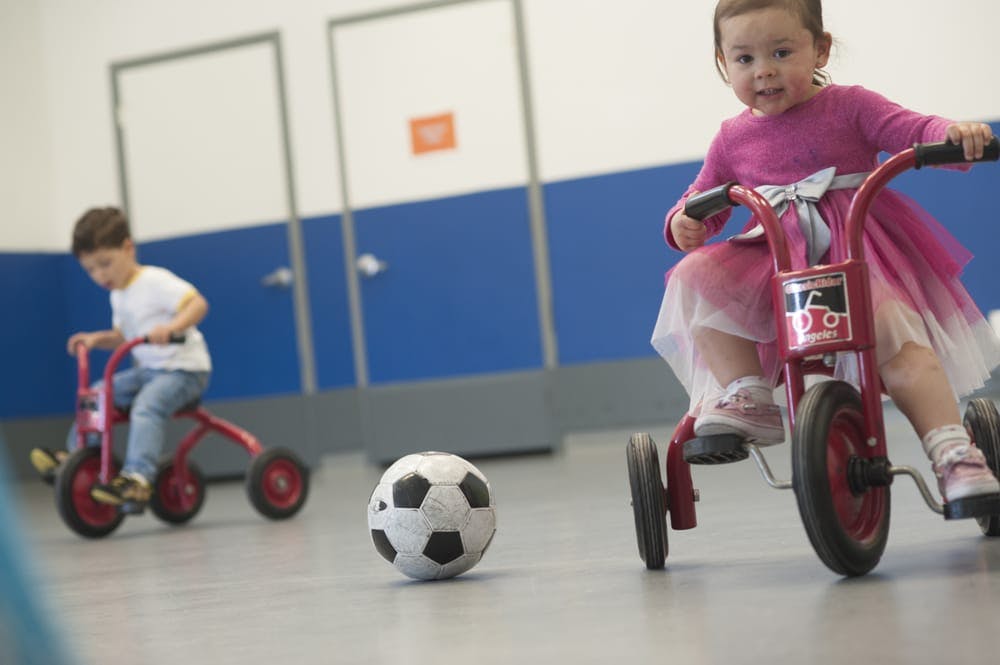 Two children riding tricycles with a soccer ball on the floor