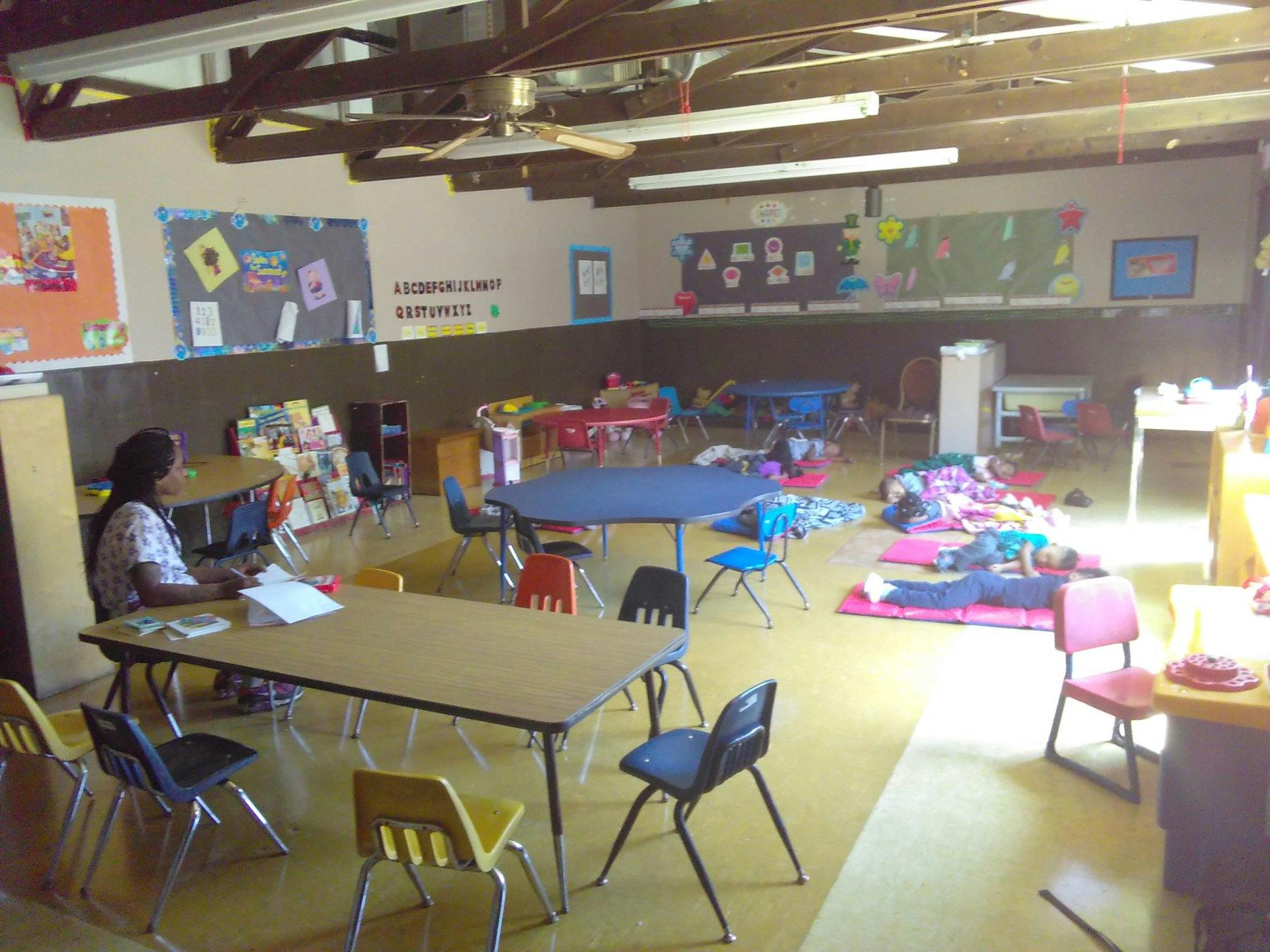 Person sitting at a table in a classroom with children sleeping on mats
