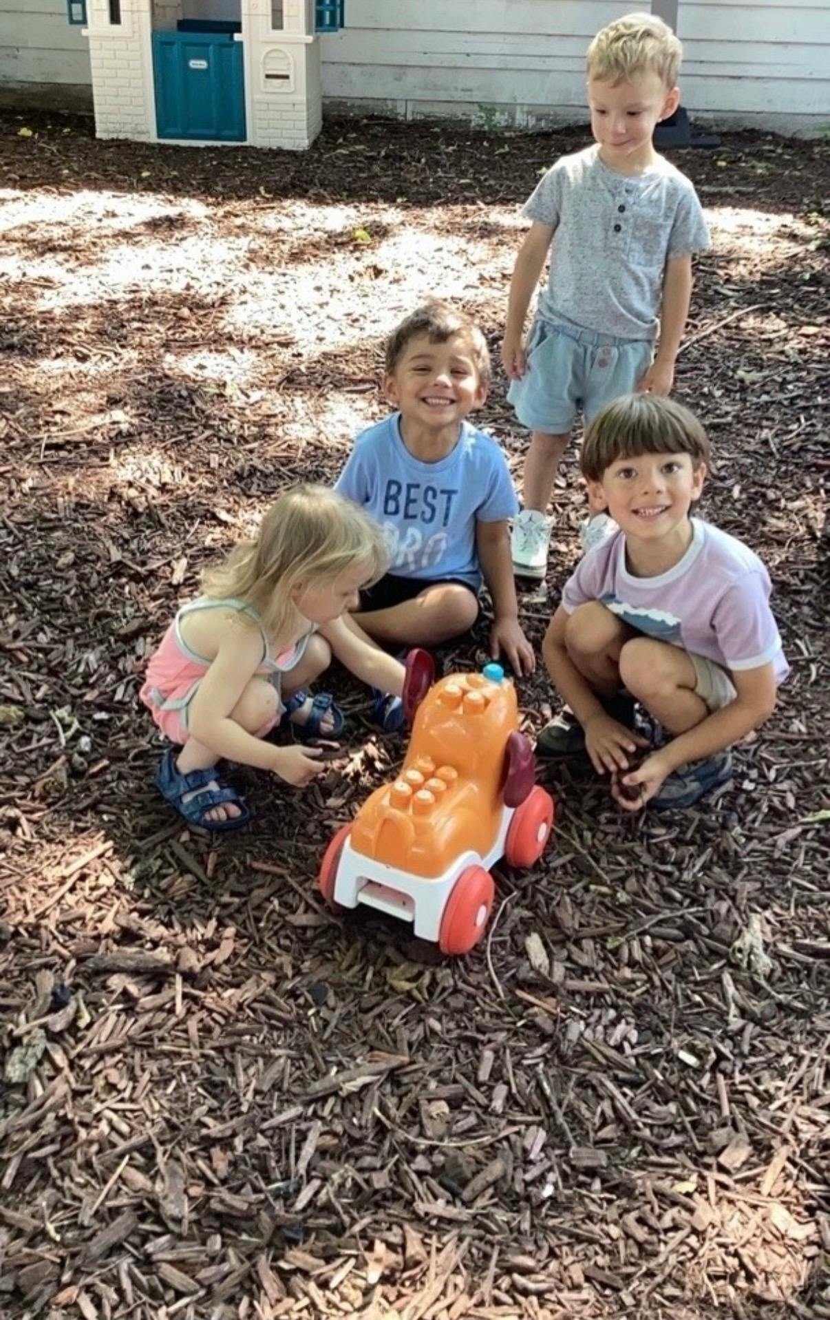 Four children playing with a toy vehicle on mulch in a yard