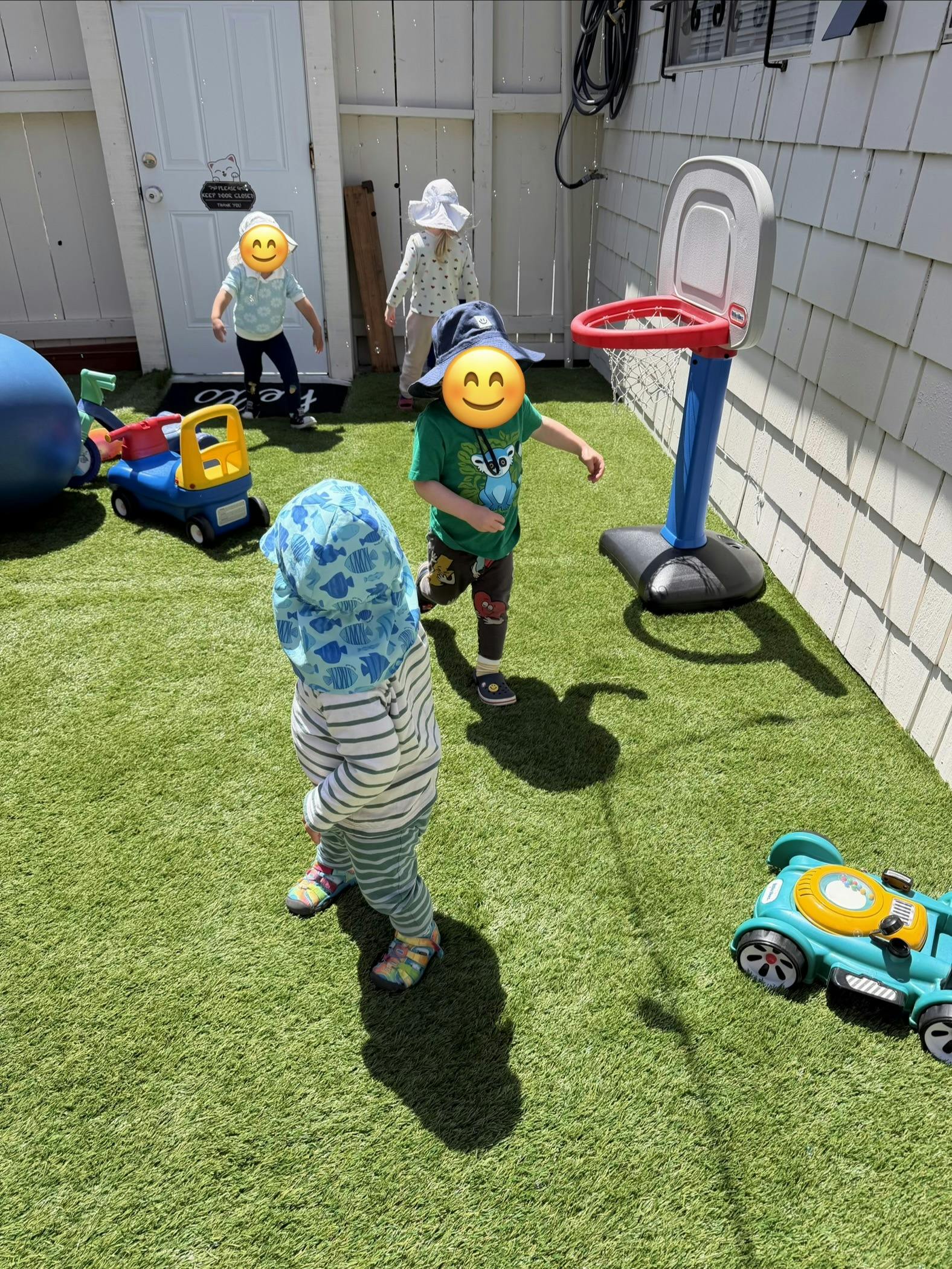 Several children in a fenced yard with toys, one reaching for a basketball hoop