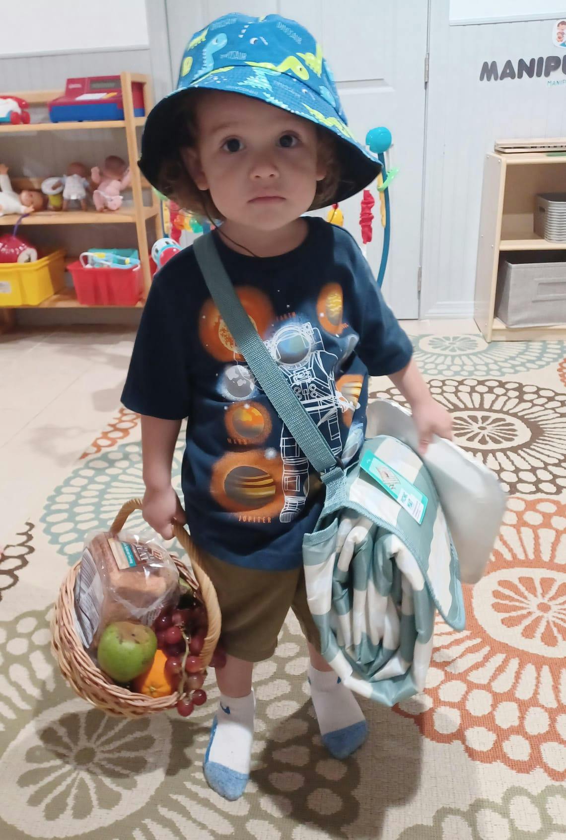 Child in a blue hat holding a basket of fruit and a rolled blanket