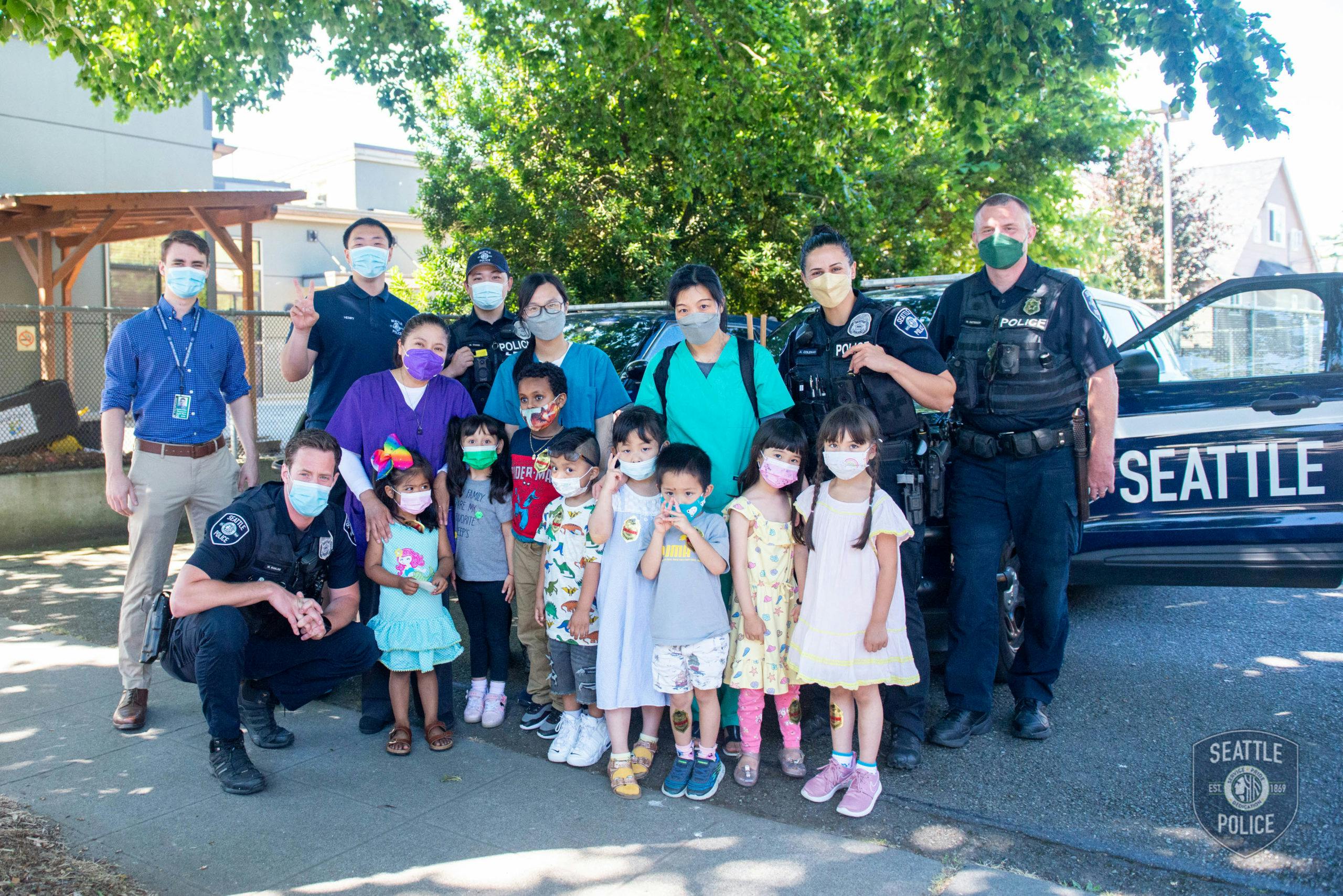 Group of adults and children wearing masks in front of a police car with "SEATTLE" written on the side
