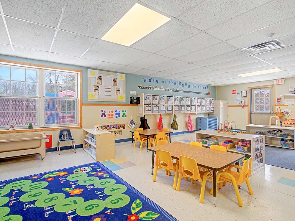 Classroom with small tables and chairs, a playmat with numbers, and toys