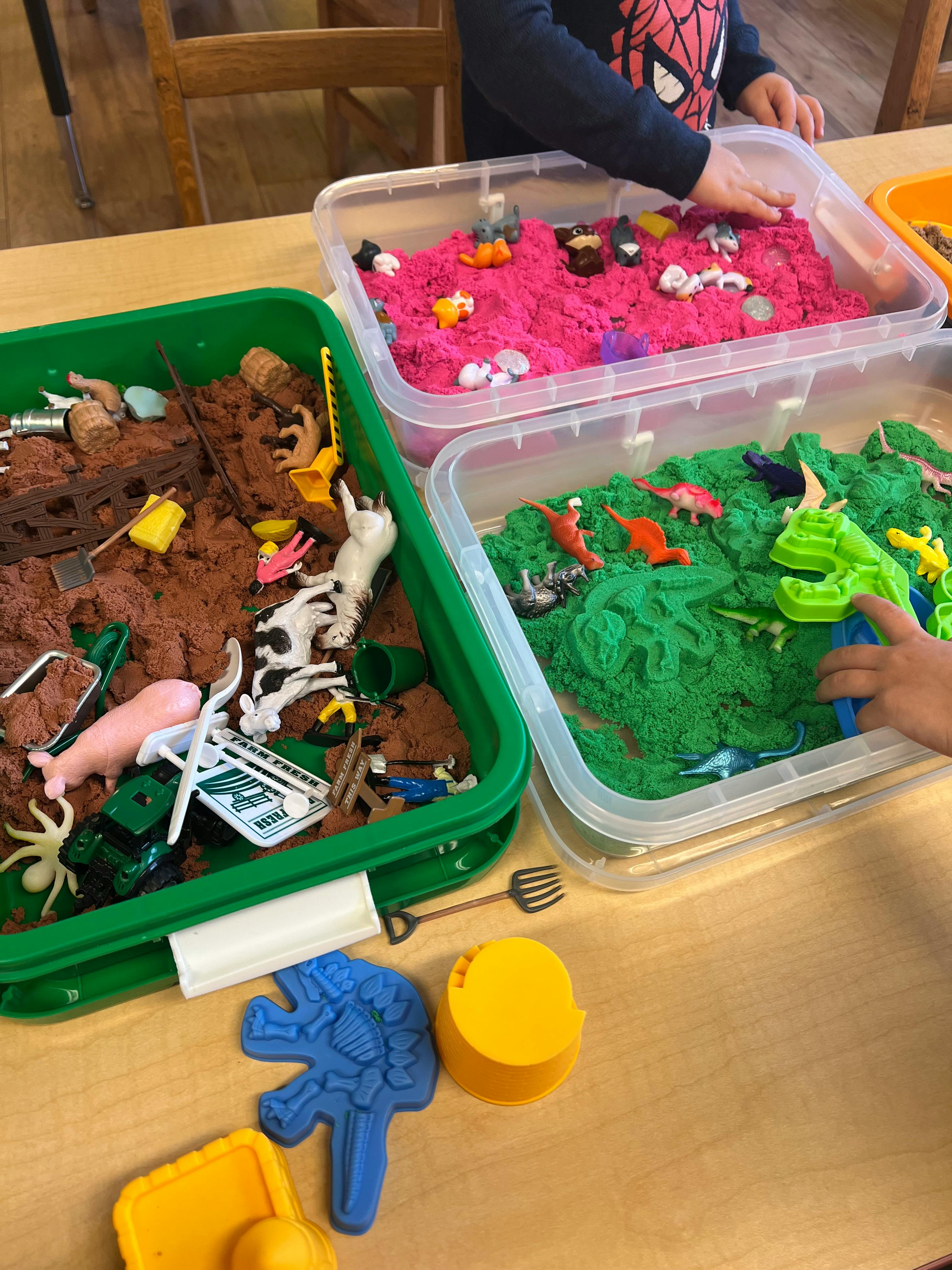 Child playing with kinetic sand and small toys in plastic bins