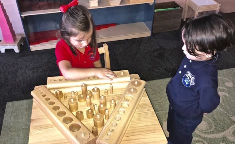 Child playing with wooden blocks at a table, another child standing nearby