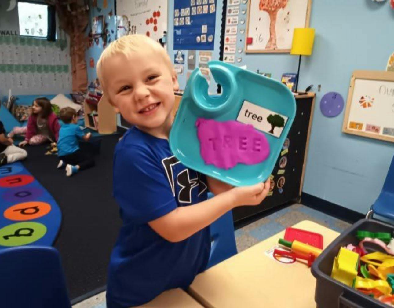 Child holding a tray with modeling clay and a tree label in a classroom