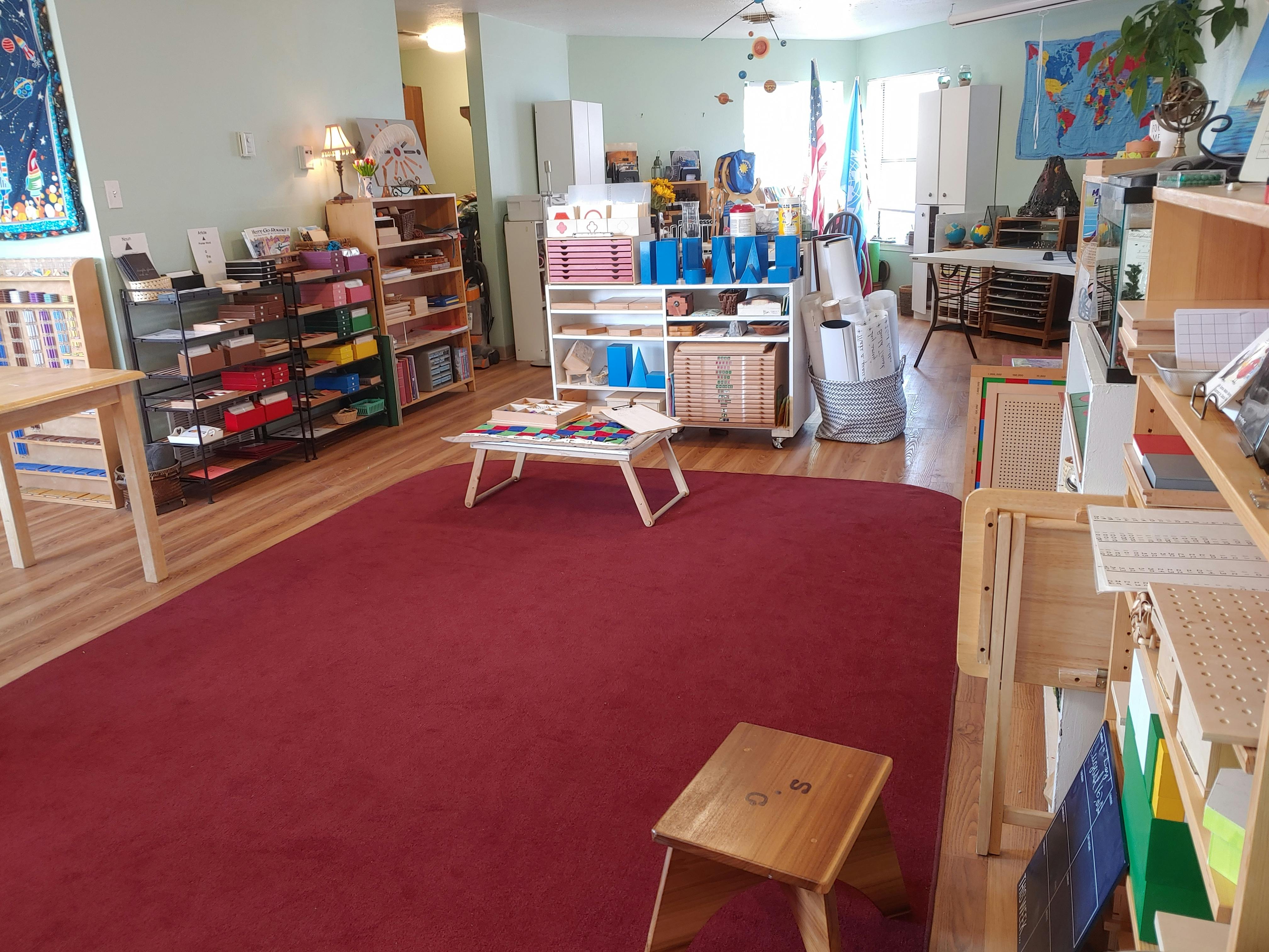 Classroom with red rug, shelves of learning materials, and small tables