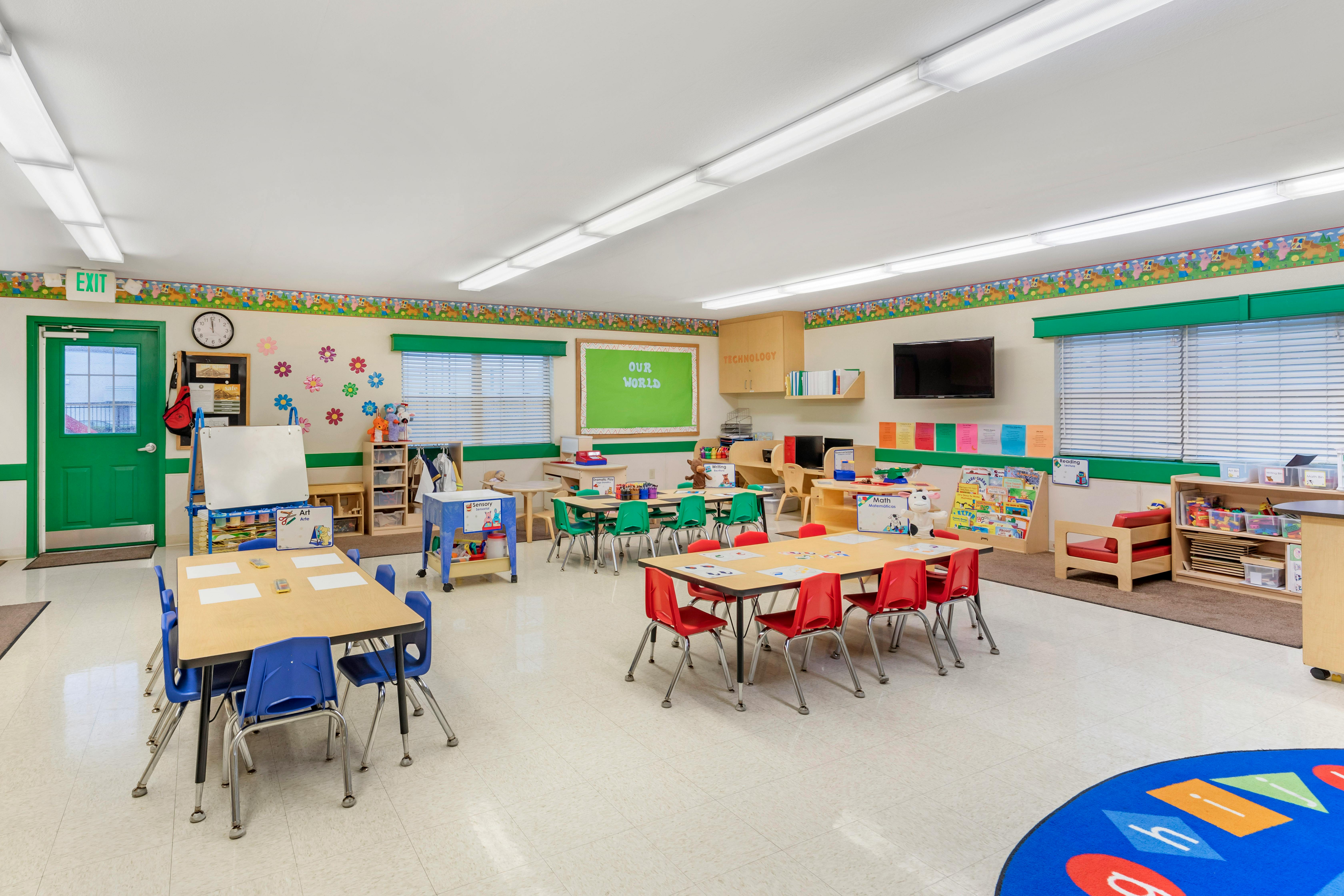 Classroom with child-sized tables and chairs, exit sign above a green door