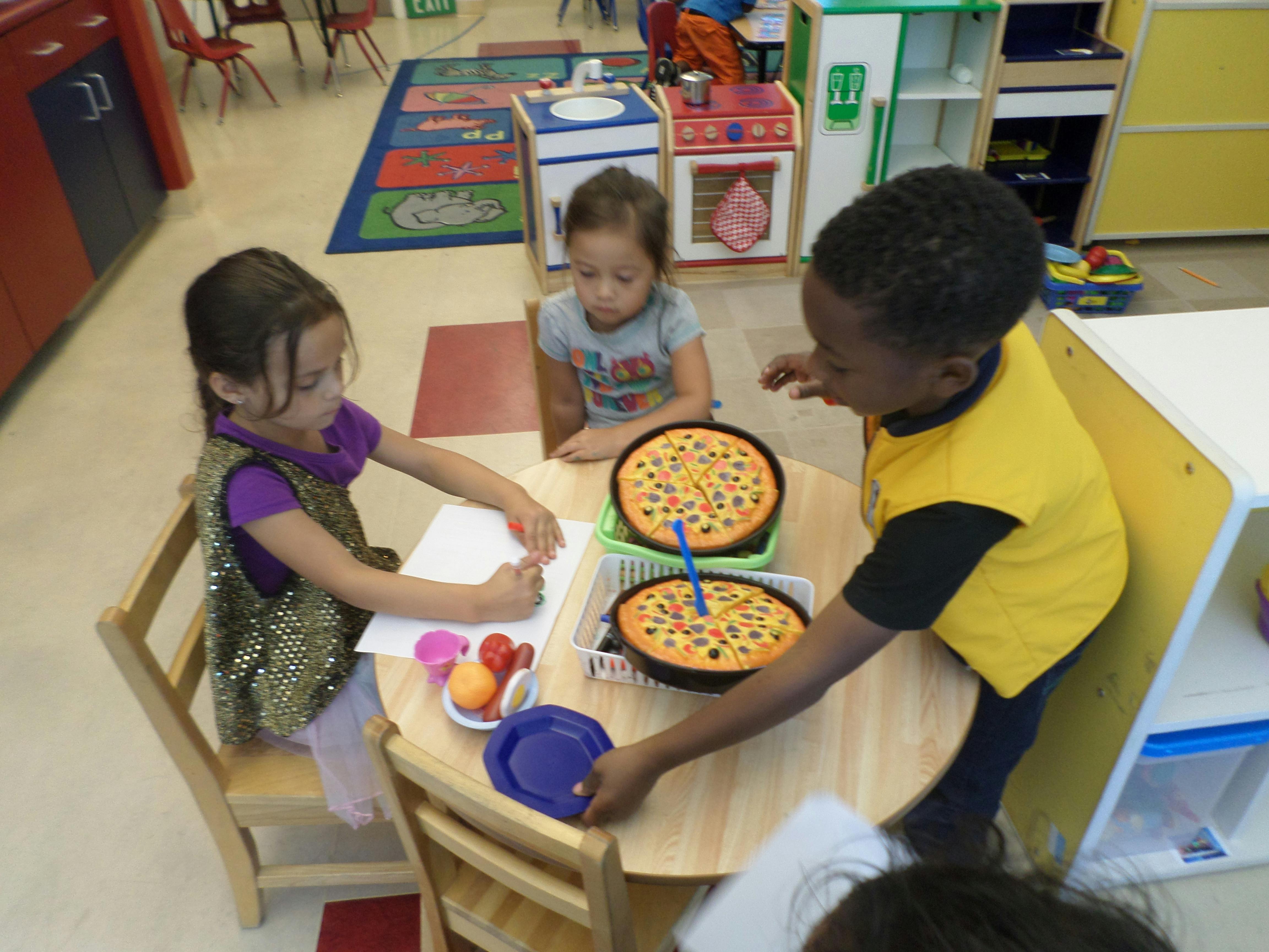 Three children playing with toy pizzas and drawing at a table