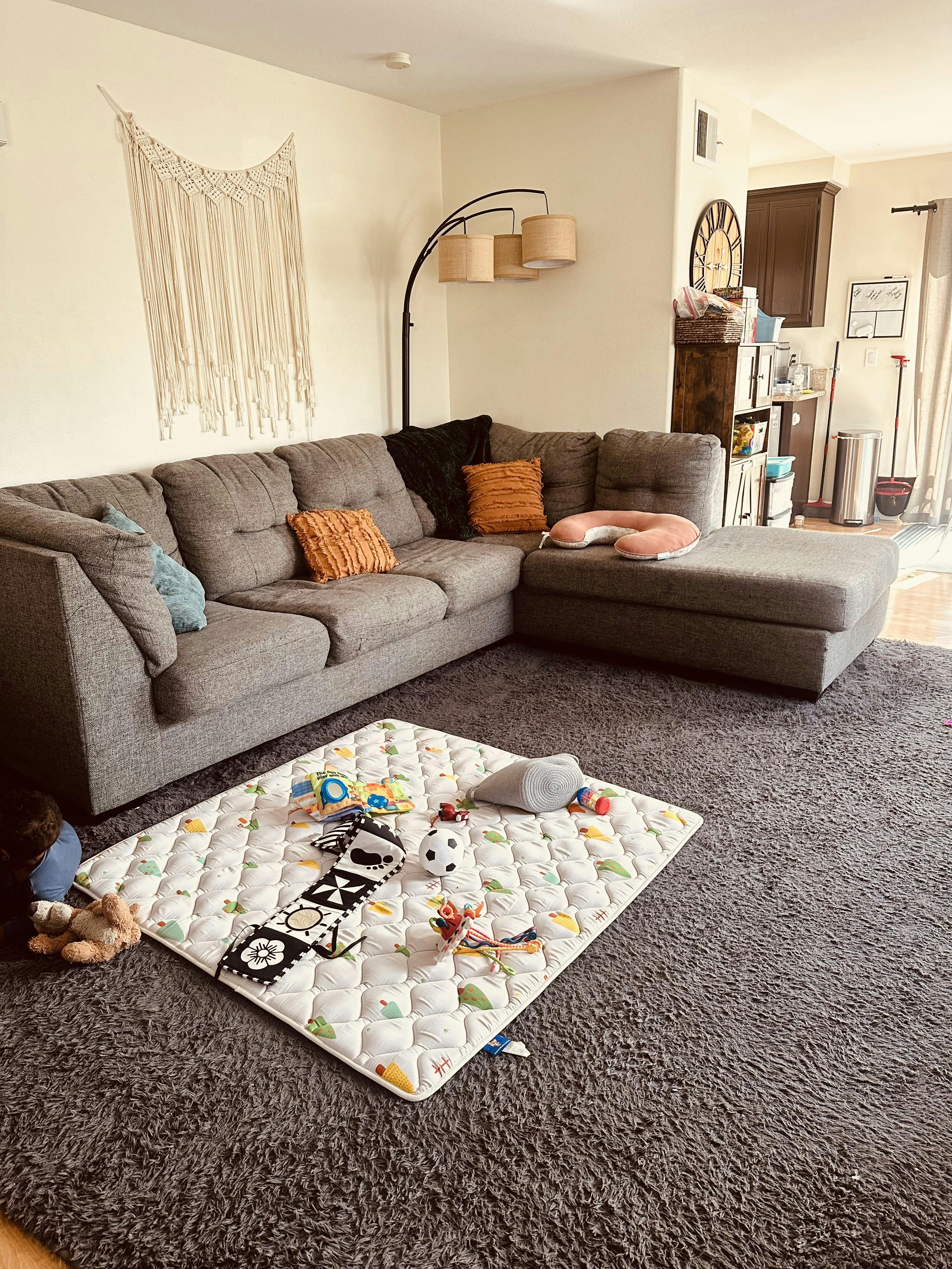 Living room with gray sectional sofa, playmat on shaggy rug, and kitchen in background