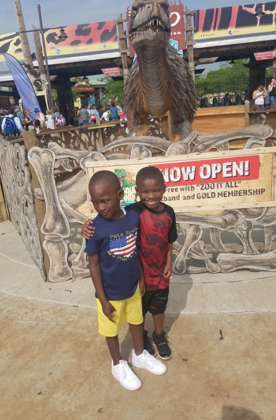 Two children standing in front of a dinosaur statue and a "NOW OPEN!" sign
