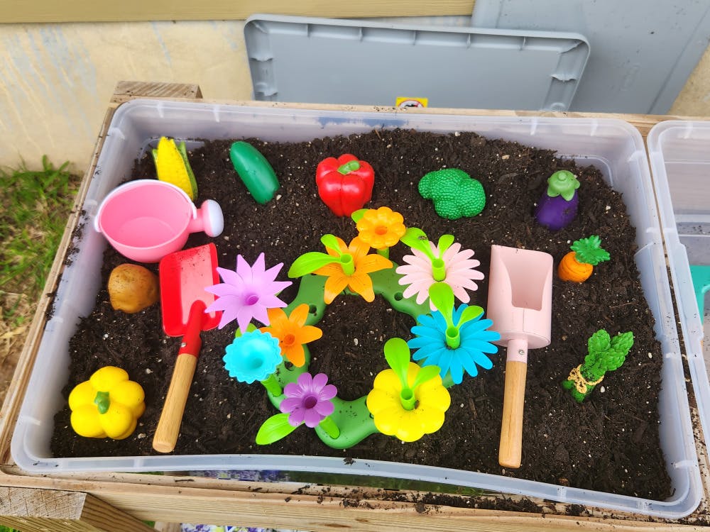 Plastic flowers, vegetables, and gardening tools in a container of soil