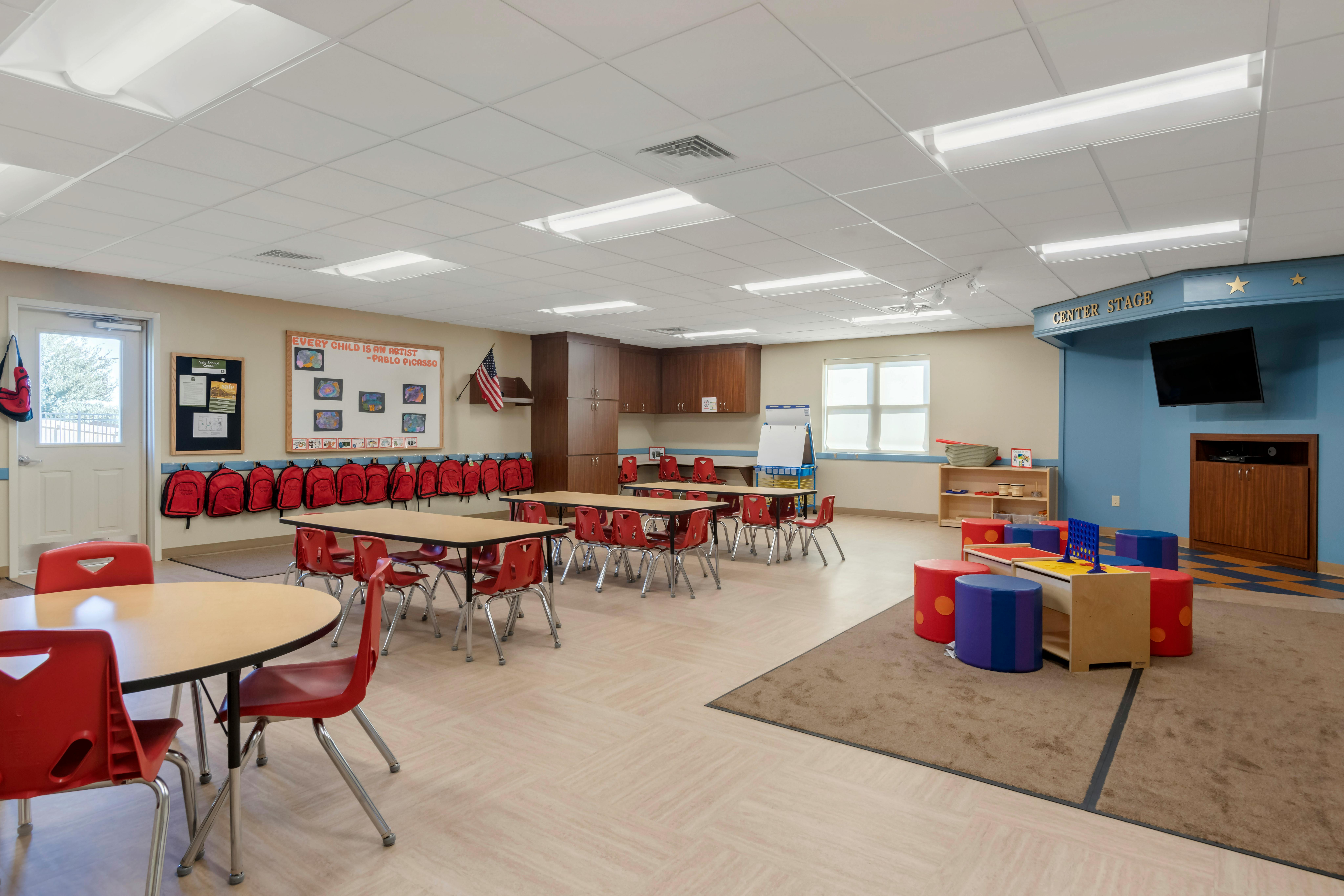 Classroom with tables, red chairs, red backpacks hanging on a wall, and a stage