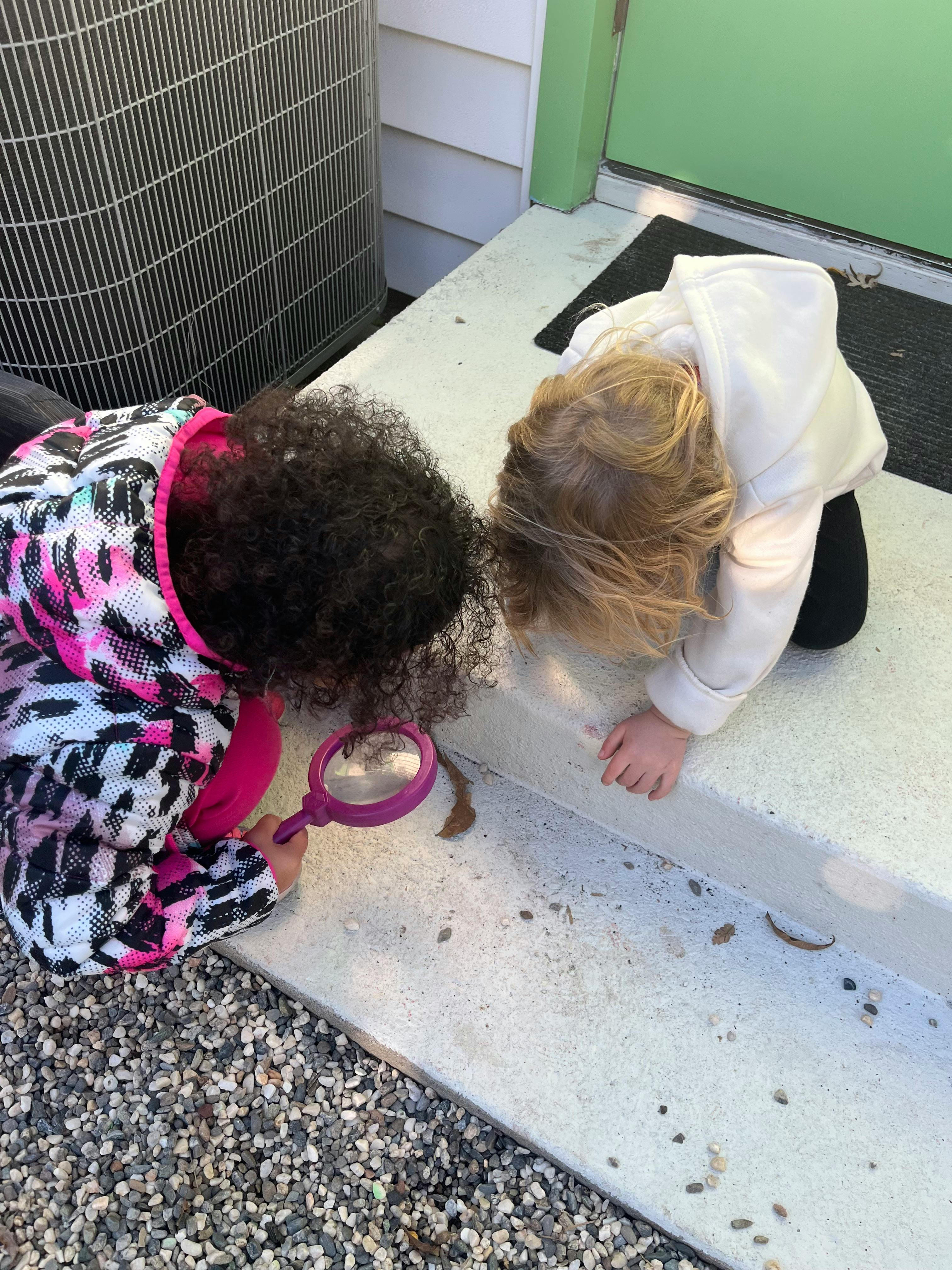 Two children looking at something on the ground with a magnifying glass