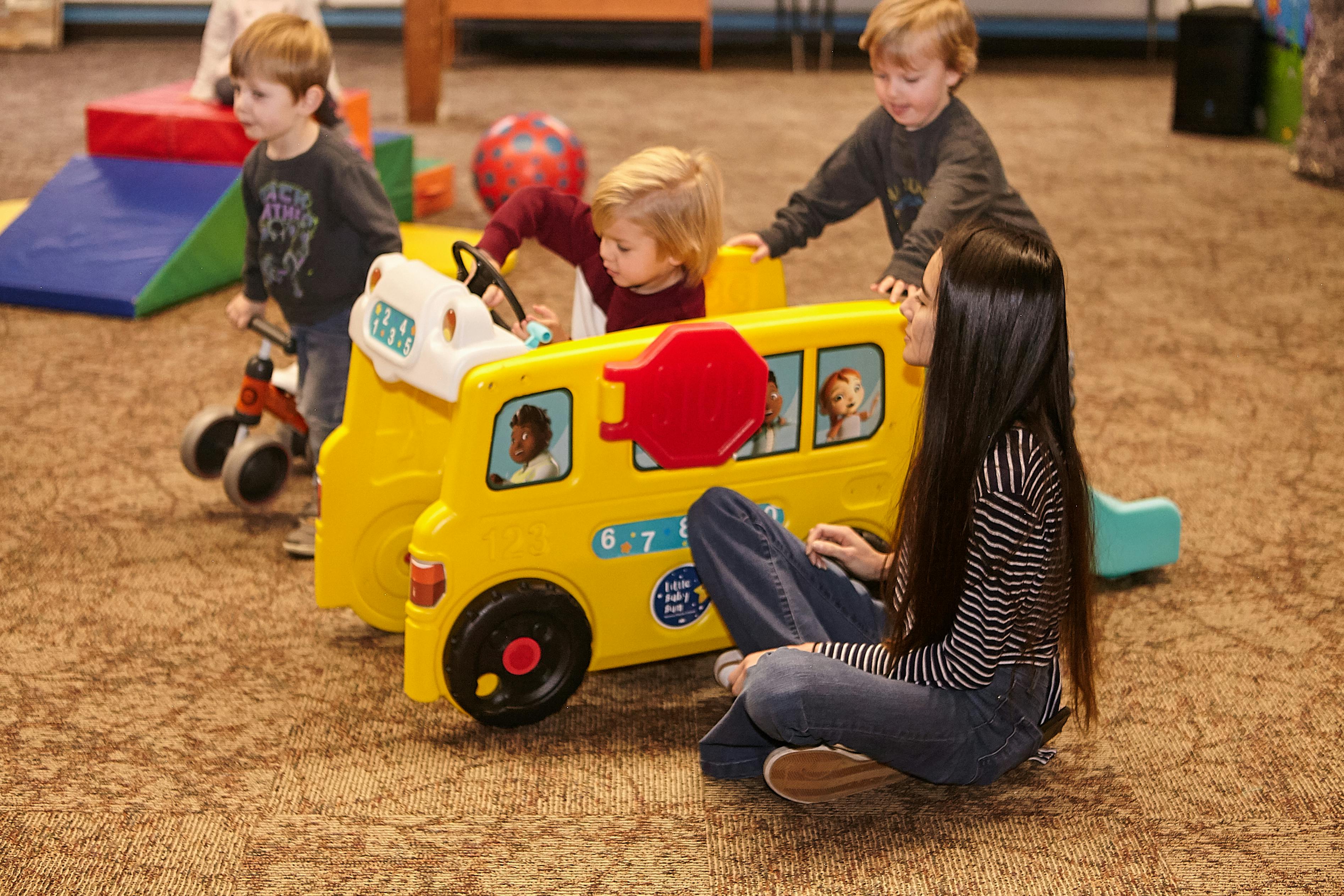 Person sitting on floor with children playing around a toy school bus