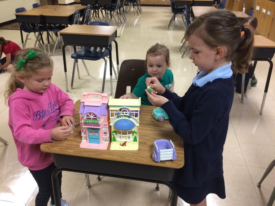 Three children playing with toy houses at a desk in a classroom